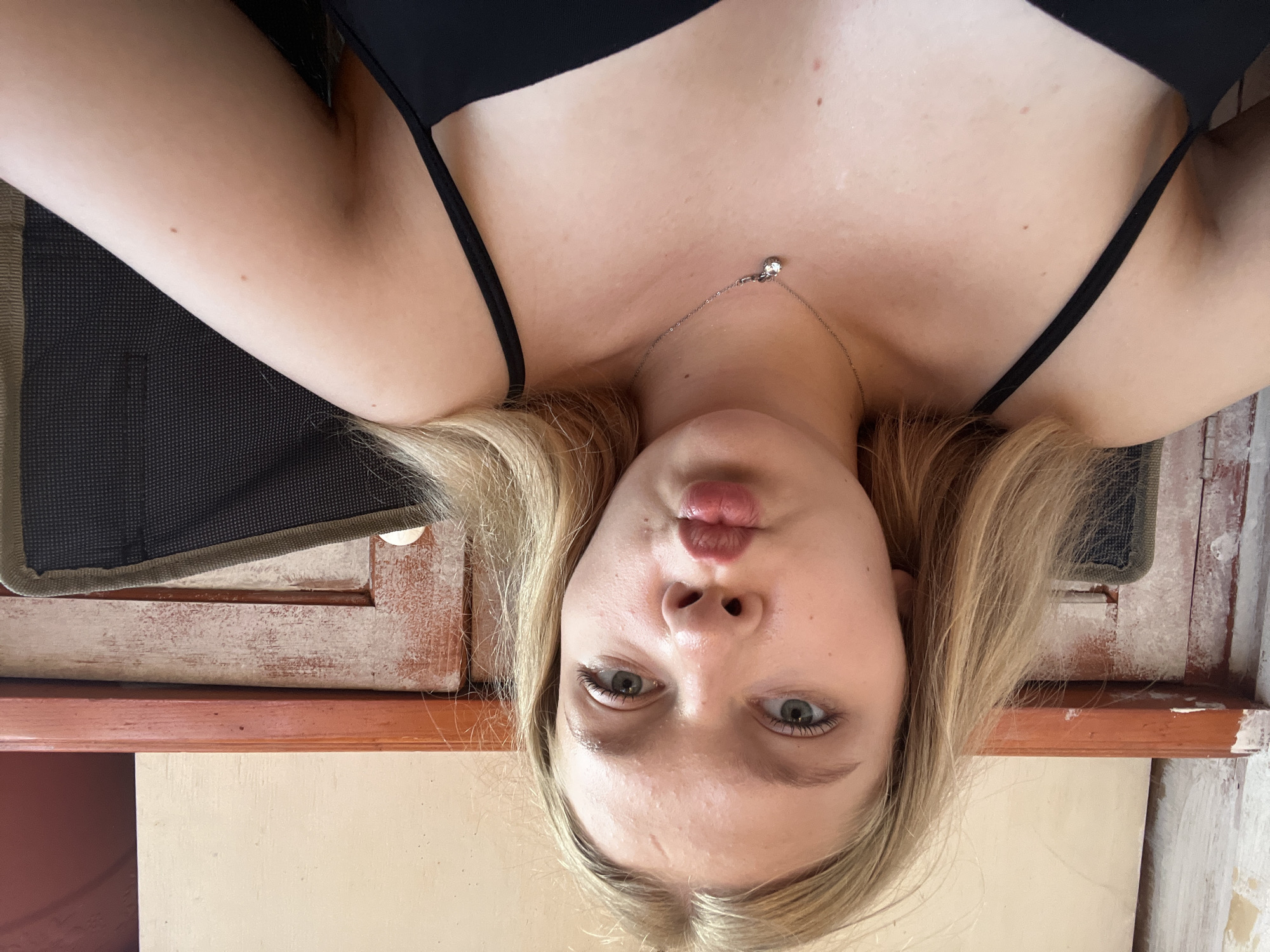A young woman with blonde hair and blue eyes is taking a selfie. She is wearing a black tank top and a silver necklace with a diamond pendant. Her lips are puckered in a kiss. The background appears to be a rustic, outdoor setting, possibly a cafe or patio, with weathered wooden elements. There are no visible landmarks to identify a specific location in Perm, Russia.