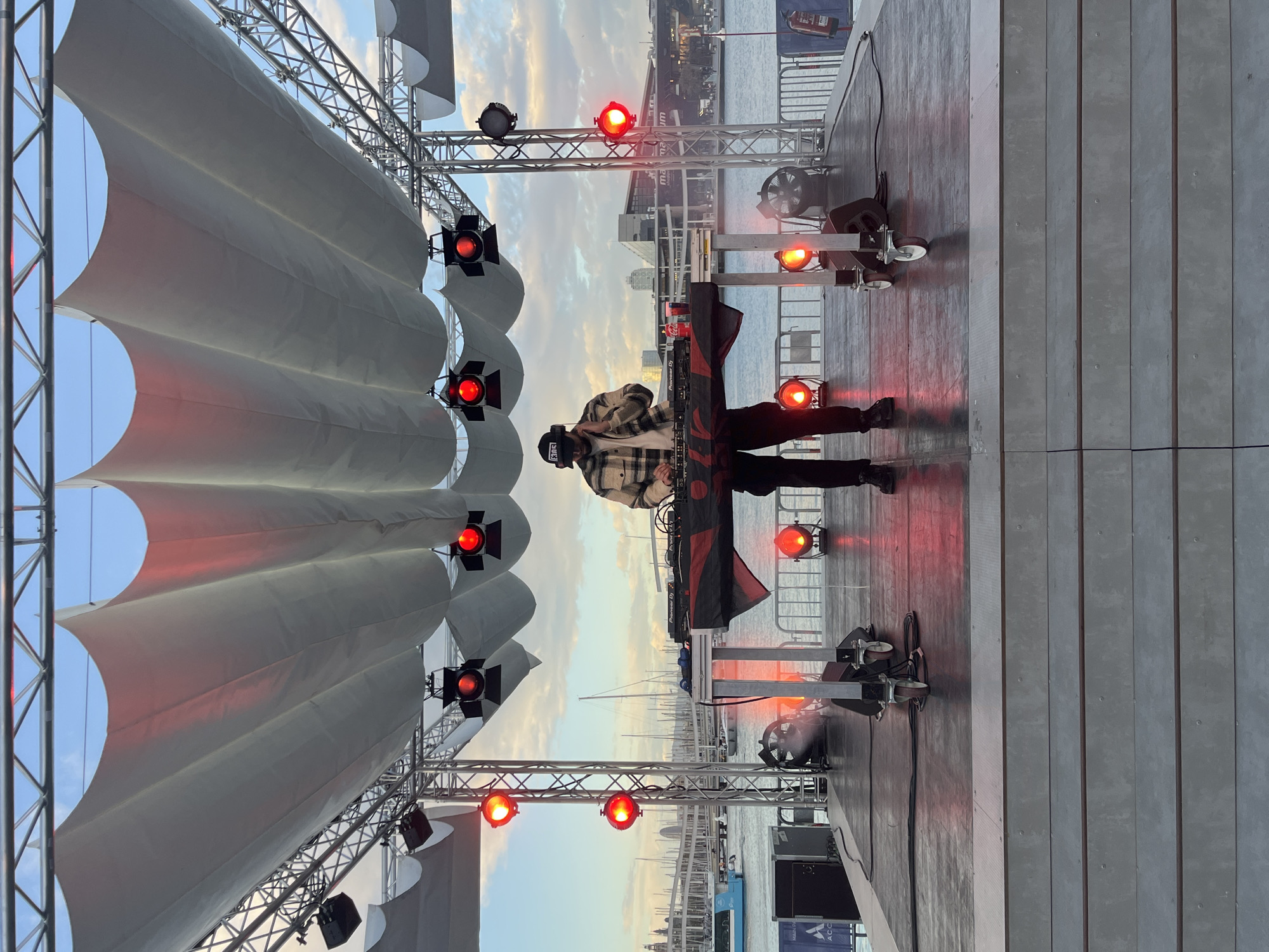 The image shows a DJ performing on an outdoor stage. The stage is covered by a white canopy supported by a metal frame, with red stage lights attached. The DJ, wearing a plaid jacket and a baseball cap, stands behind a Pioneer DJ mixer. The background reveals a cityscape and a body of water, possibly a harbor. Based on the architecture and the presence of water, the location is likely Barcelona, Spain.