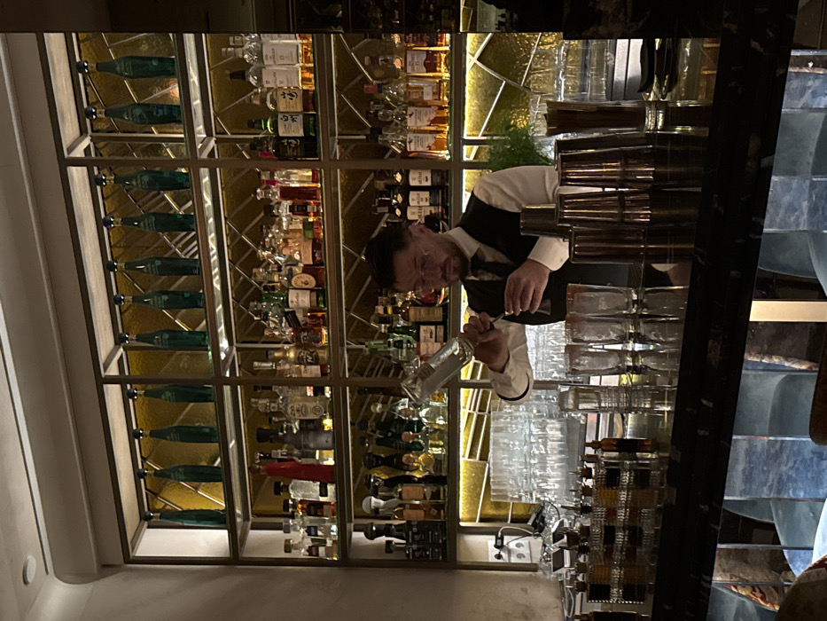 A bartender in a vest and white shirt is pouring liquid from a clear bottle. The setting is a bar, with multiple shelves displaying rows of liquor bottles against a gold-toned backdrop. Glasses are stacked in front of the shelves. The image is slightly tilted, showing parts of the ceiling and wall on the left, and a blue-toned textured surface on the right.