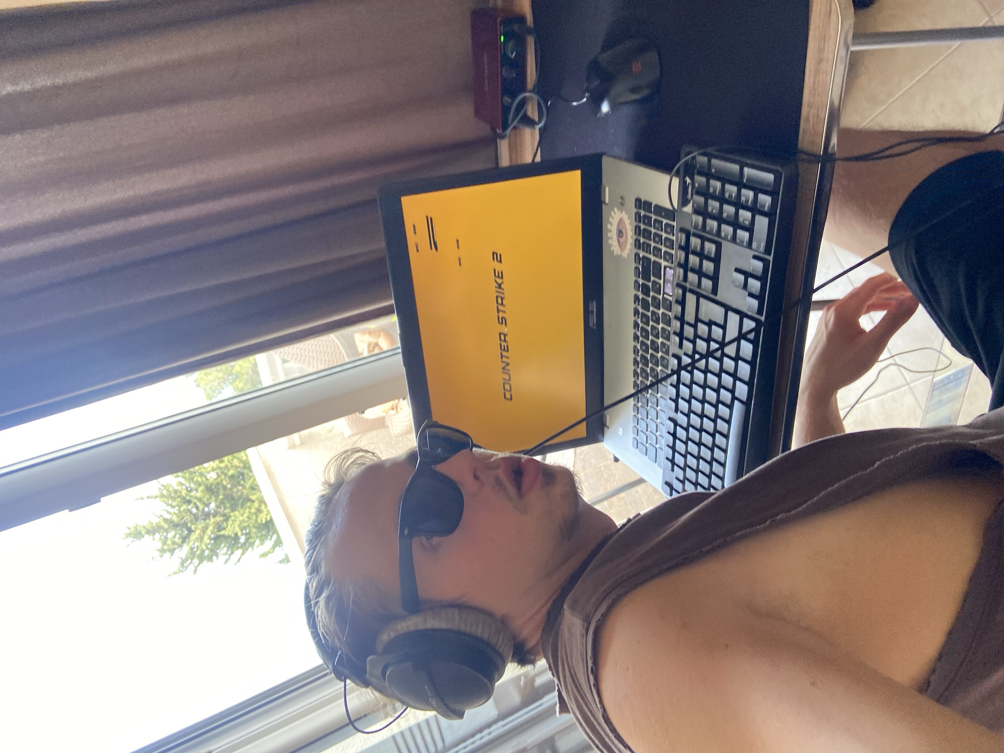 A man with sunglasses and headphones sits at a desk, playing Counter-Strike 2 on a laptop. A mouse and keyboard are also on the desk. The man is in a room with a window overlooking trees and a drawn curtain. The location is not definitively identifiable as Šušanj, Montenegro.