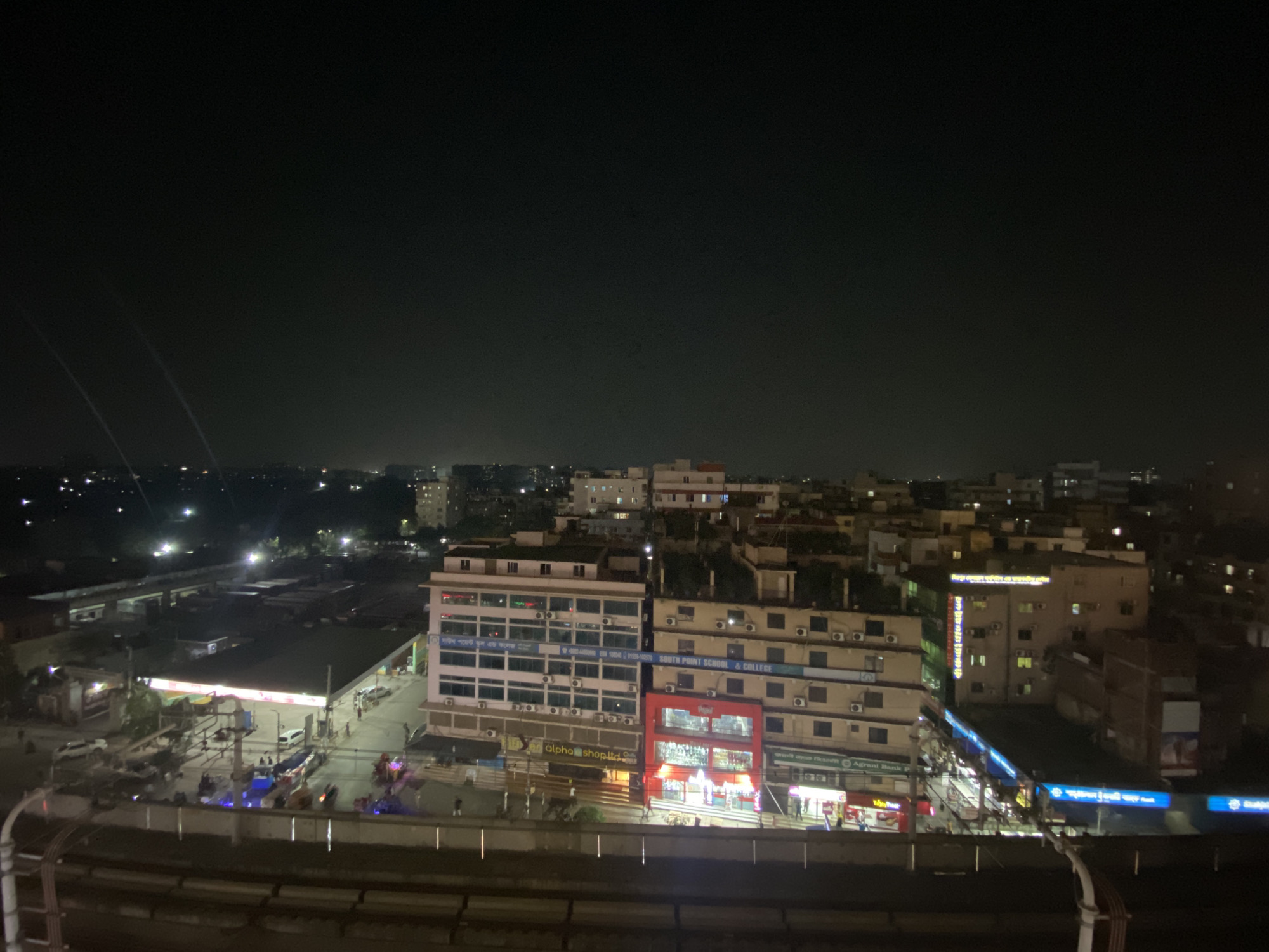 The image depicts a nighttime scene in Dhaka, Bangladesh, showcasing an urban area with buildings, streetlights, and a railway line in the foreground. The "South Point School & College" signage is visible, indicating the location. In the foreground is a railway track with lighting, and further away, the buildings are clustered. Street-level businesses and shops are lit up, indicating activity in the area. People are present but appear small and indistinct due to the distance and darkness. The overall environment suggests an active city life continuing after dark.