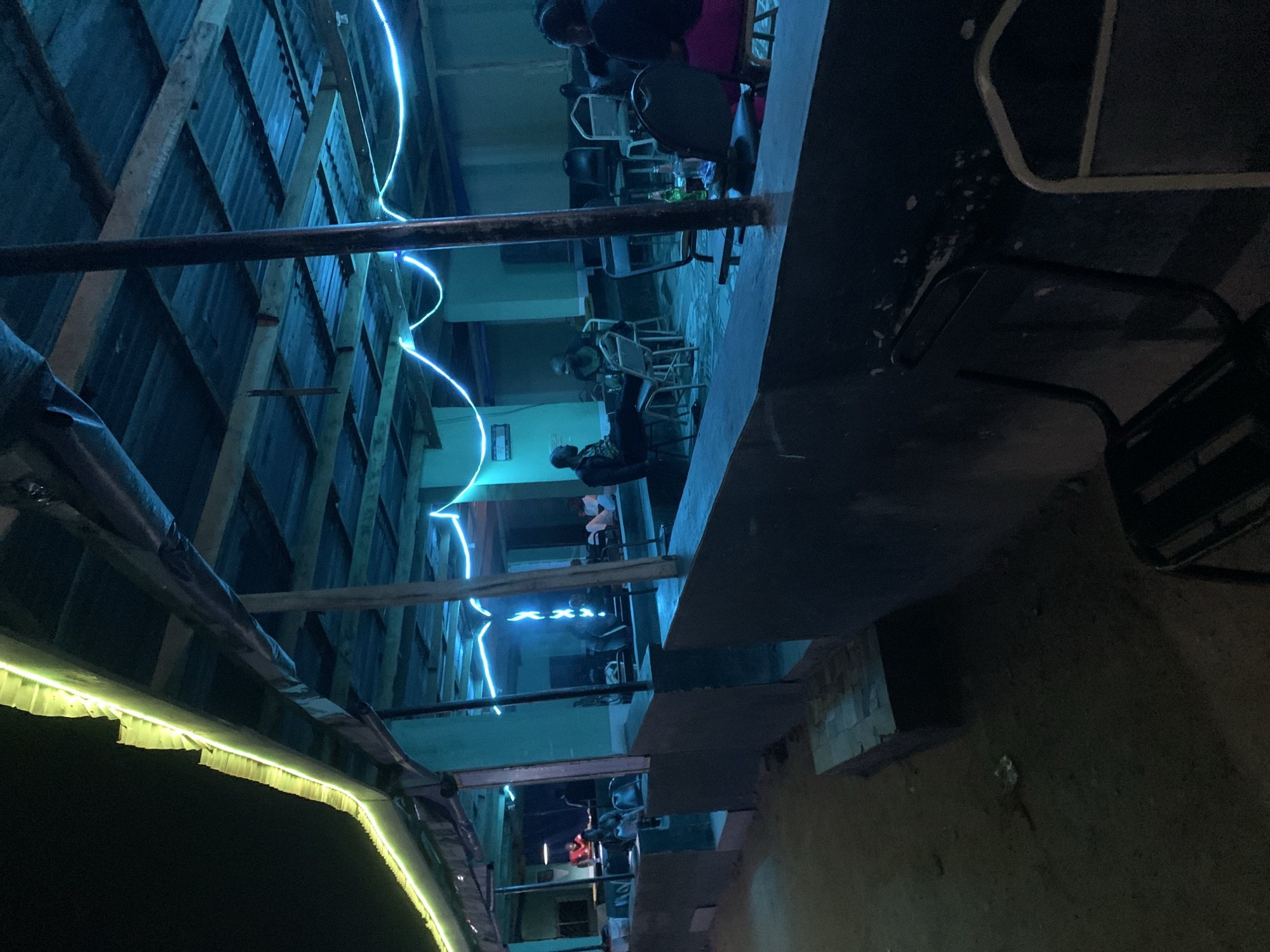 The image shows a view looking down an alley or outdoor seating area in New Stadium, Nigeria. Patrons are seated at tables and chairs, likely enjoying drinks or food. Blue and yellow decorative lights illuminate the space. The alley appears constructed with corrugated metal sheets and wooden supports. The overall atmosphere seems dim, with the lights providing the primary source of illumination.