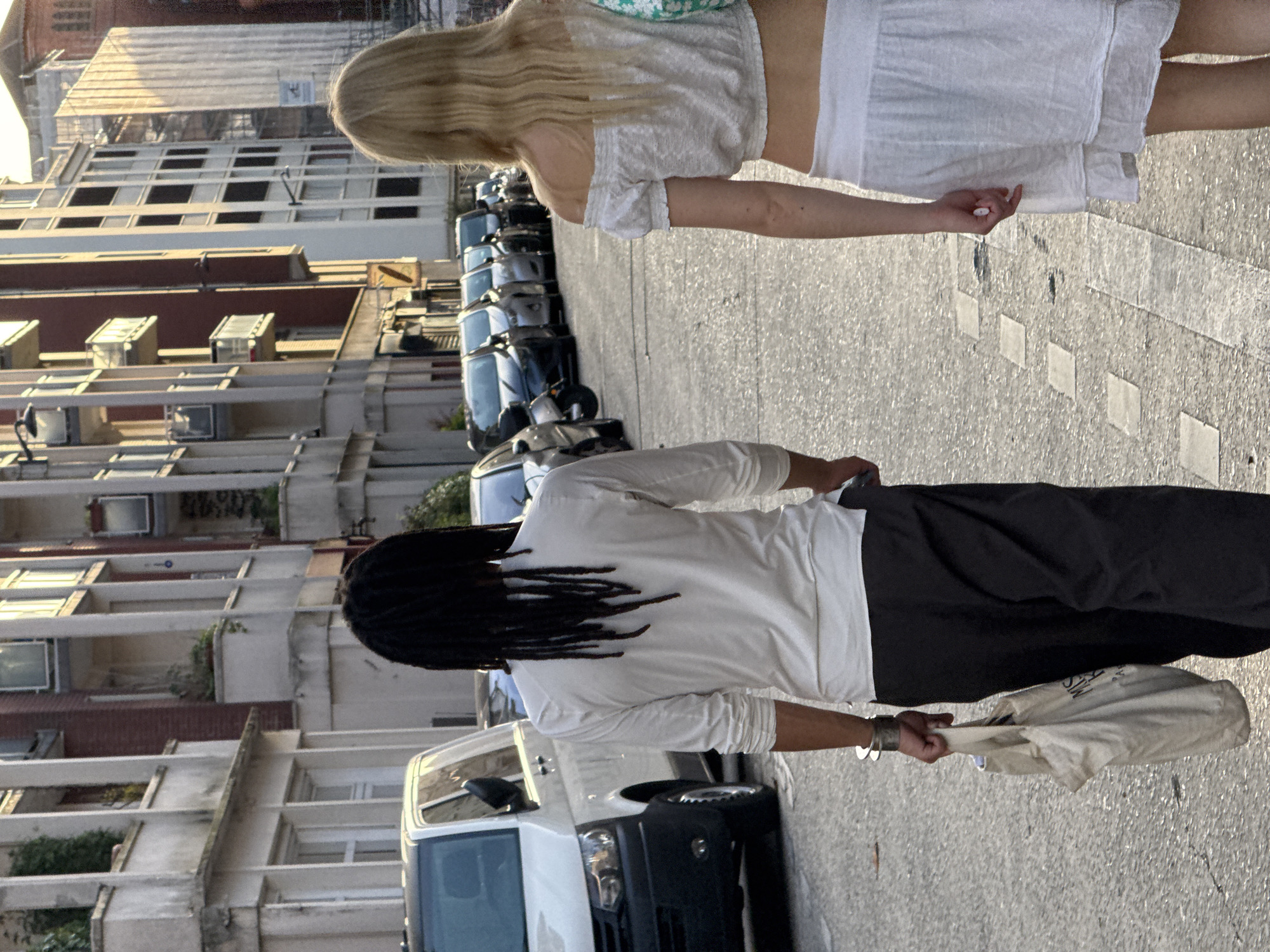 A medium shot shows two women walking on a street. The woman in the foreground is wearing a white long-sleeve shirt and dark pants. She also has dreadlocks and carries a cream-colored bag. The other woman is wearing a white crop top and a white skirt. They are both walking on the right side of the road. Cars are parked on the left side of the road, and a van is directly behind the woman in the foreground. Tall buildings line the street. This scene could be in Paris, France.