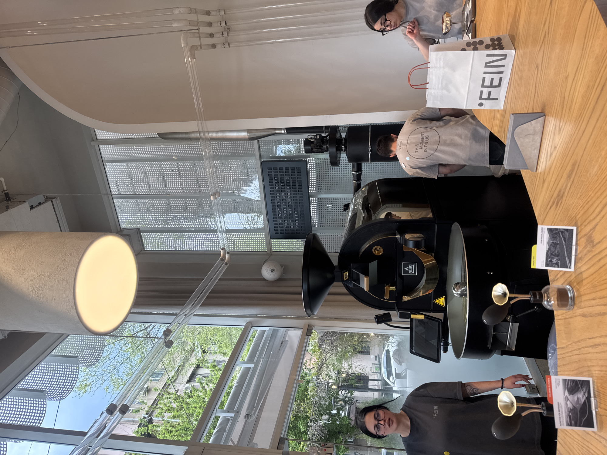The image captures the interior of a modern coffee roastery or cafe in Yerevan, Armenia, characterized by a clean, minimalist aesthetic and ample natural light. The scene appears to be active during daytime, with bright sunlight streaming through large windows that offer a view of lush green trees and distant buildings.
Three individuals are visible. In the foreground, on the left, a woman wearing glasses and a dark gray t-shirt with the word "FEIN" printed on it stands behind a light wooden counter, looking towards the viewer. To the right of the central coffee roasting machine, another person, with their back partially turned, wears a light beige t-shirt that reads "THIS MOMENT IS ON US." Further to the upper right, a third person, also wearing glasses and a light-colored t-shirt, stands facing the counter, possibly engaging with another person or examining products.
The central object is a prominent, large black coffee roasting machine, branded "PROBAT," suggesting active coffee roasting operations are a key part of this establishment. On the wooden counter, next to the roaster, are two small glass containers with brass-colored elements, likely used for presenting coffee beans or grounds for aroma sampling. Adjacent to these are small information cards, one clearly stating "FEIN SPECIAL BLEND" and another "ROASTED FOR ESPRESSO." A white paper shopping bag with the "FEIN" logo is also on the counter.
The interior design features transparent tubes mounted on the ceiling and walls, contributing to the contemporary feel. A large, cylindrical light fixture hangs from the ceiling. In the background, behind the roaster, a black letter board displays the quirky message: "I'M NOT SURE WHAT THE QUESTION IS BUT THE ANSWER IS COFFEE." The overall impression is that of a specialized coffee shop or roastery, focusing on high-quality coffee and a modern customer experience.
