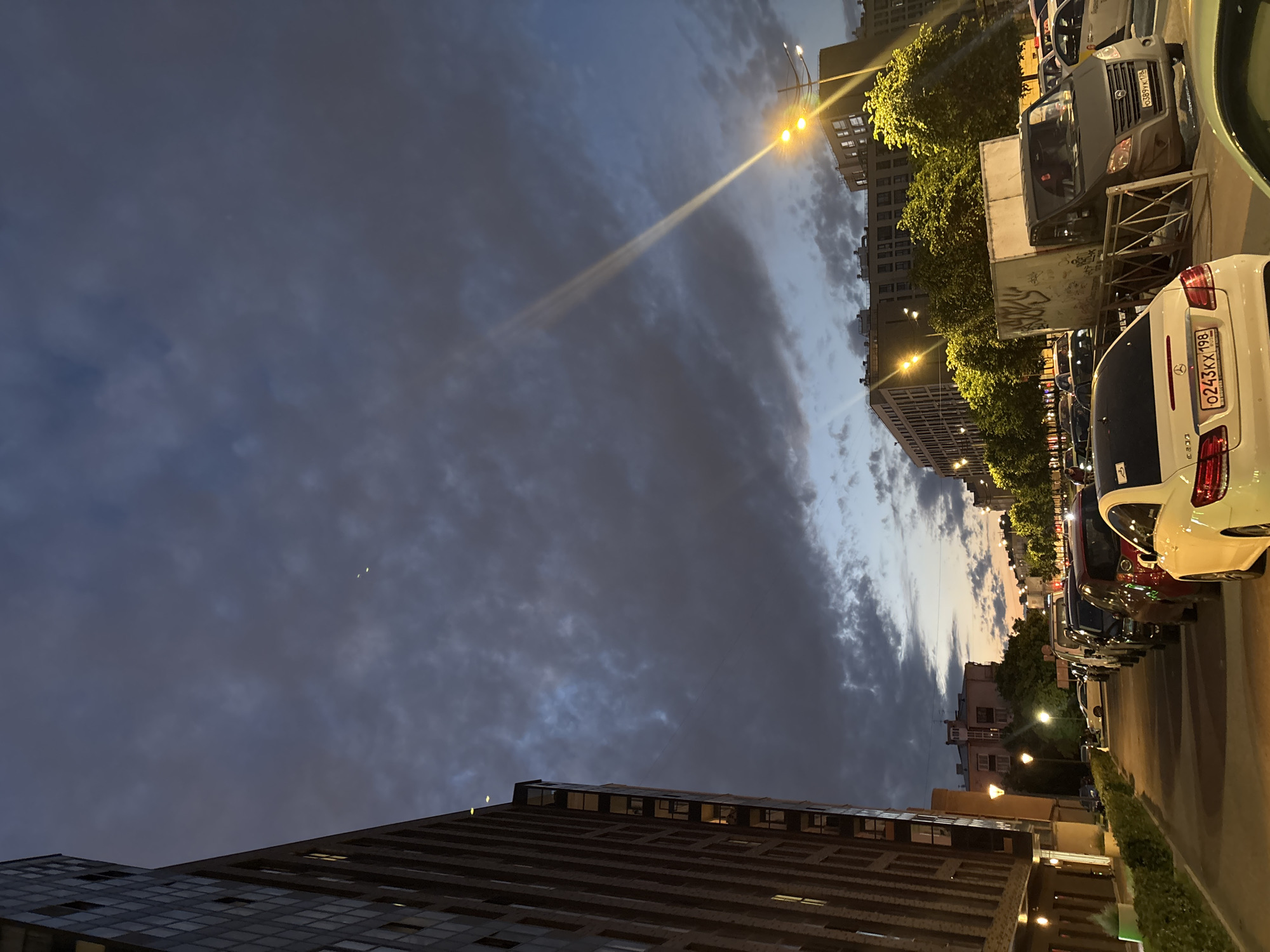 This is a photograph taken at street level in the city of Saint Petersburg, Russia, during twilight. The image features a street lined with parked cars and modern buildings. On the left, a tall, multi-story brick building with many windows dominates the foreground. Opposite this building, a row of parked cars extends down the street. The sky above is a mix of dark, cloudy formations and patches of lighter blue and orange hues, suggesting the last moments of daylight. Streetlights are visible, casting yellow illumination on the scene. A white car with a license plate ending in "198" is parked prominently in the foreground. A light-colored truck with graffiti on its side is also visible in the middle ground. The overall impression is an urban scene at dusk, with a focus on the architecture, vehicles, and the atmospheric sky.