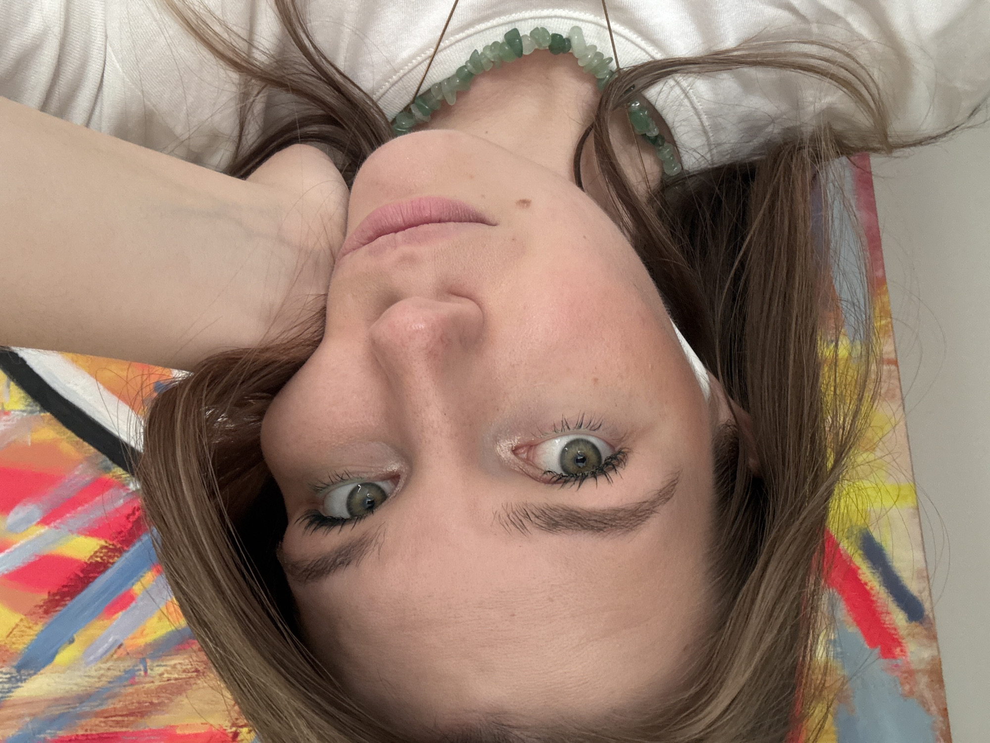 An inverted close-up portrait shows a young woman lying down and looking directly at the camera. Her head is positioned upside down within the frame, with her light brown hair spread out around her. She wears a plain white long-sleeved top and a chunky green beaded necklace made of irregular shaped stones. Her right arm is bent, with her hand resting lightly near her chin.
Her eyes, a striking light green or hazel, are wide and focused on the viewer, framed by dark eyelashes. Her eyebrows are natural and defined. Her complexion appears clear, and her lips are a soft pink. A small mole is visible on her right cheek.
Behind her head and to the viewer's left, a colorful, abstract painting is partially visible. It features bold, energetic brushstrokes in shades of red, yellow, blue, black, and white, adding a vibrant artistic element to the background. The image conveys a direct and contemplative gaze amidst a colorful setting. No text is visible.