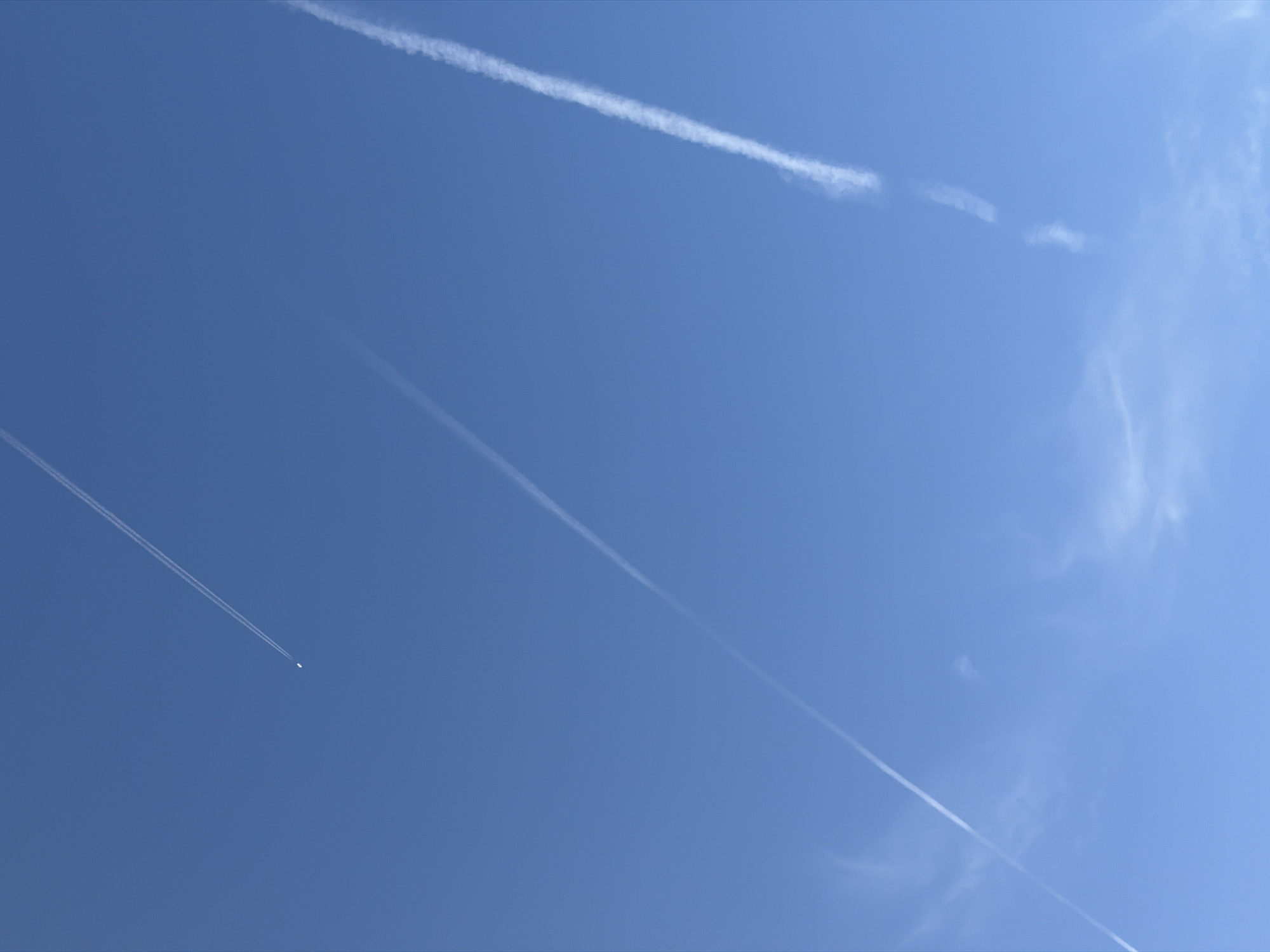 The image displays a clear blue sky with three distinct contrails from airplanes. The largest and most prominent contrail runs diagonally from the bottom right towards the upper center of the frame, appearing somewhat broken and wispy. To the left of this, another contrail extends from the bottom left towards the upper left, also appearing as a thin white line. In the upper portion of the image, a third contrail is visible, originating from a small, distant white object that is likely an airplane, and extending towards the top. There are no visible landmarks or structures suggesting a specific location in the city of Leudelange, Luxembourg. The scene is devoid of people or any other discernible subjects. The environment is solely the sky.