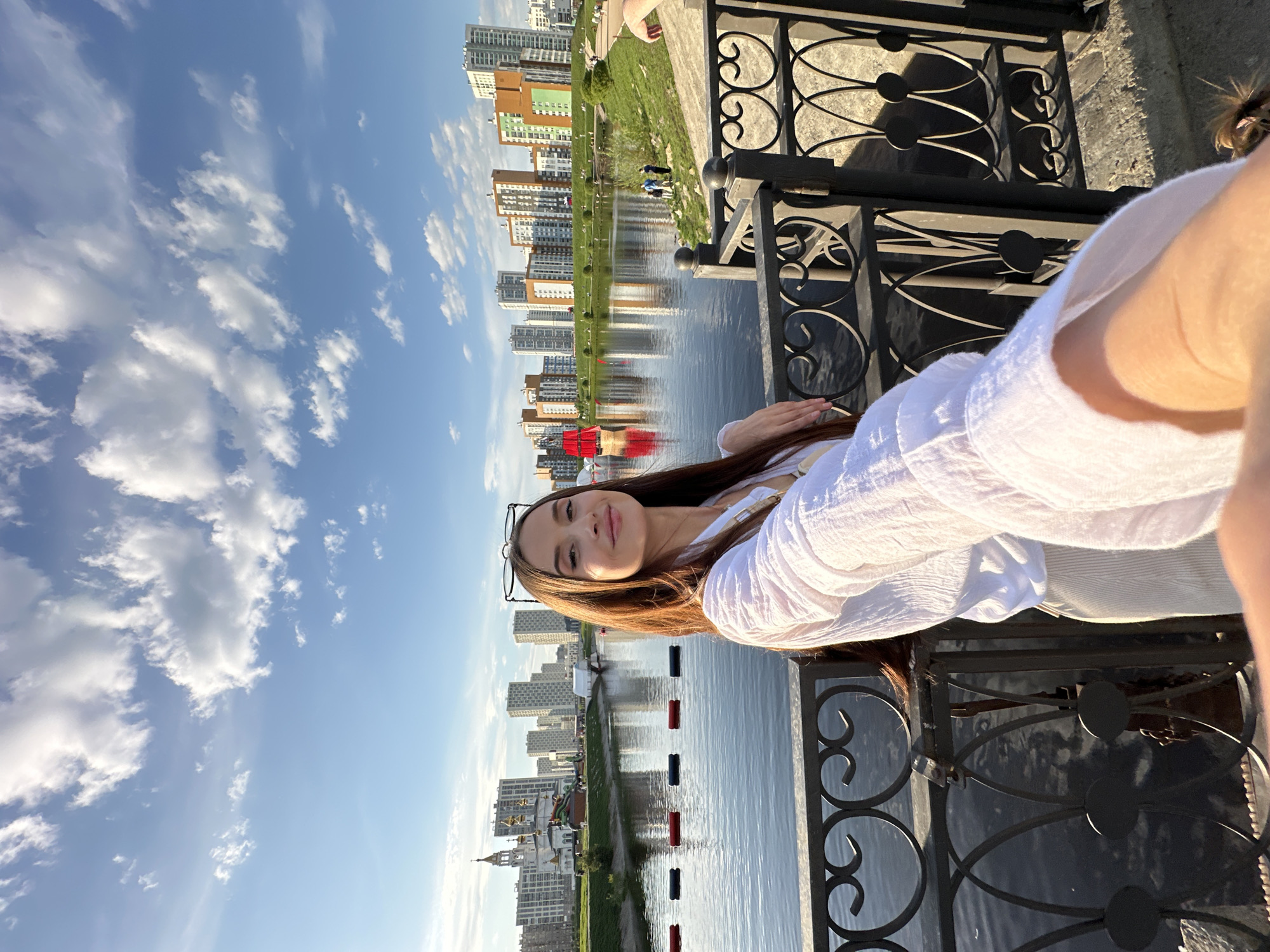 A young woman in a white blouse and glasses stands on a bridge, looking into the camera for a selfie, in Yekaterinburg, Russia. The bridge has a black metal railing with decorative swirls. Behind her is a body of water reflecting the buildings of Yekaterinburg. The sky is bright blue with puffy white clouds.
