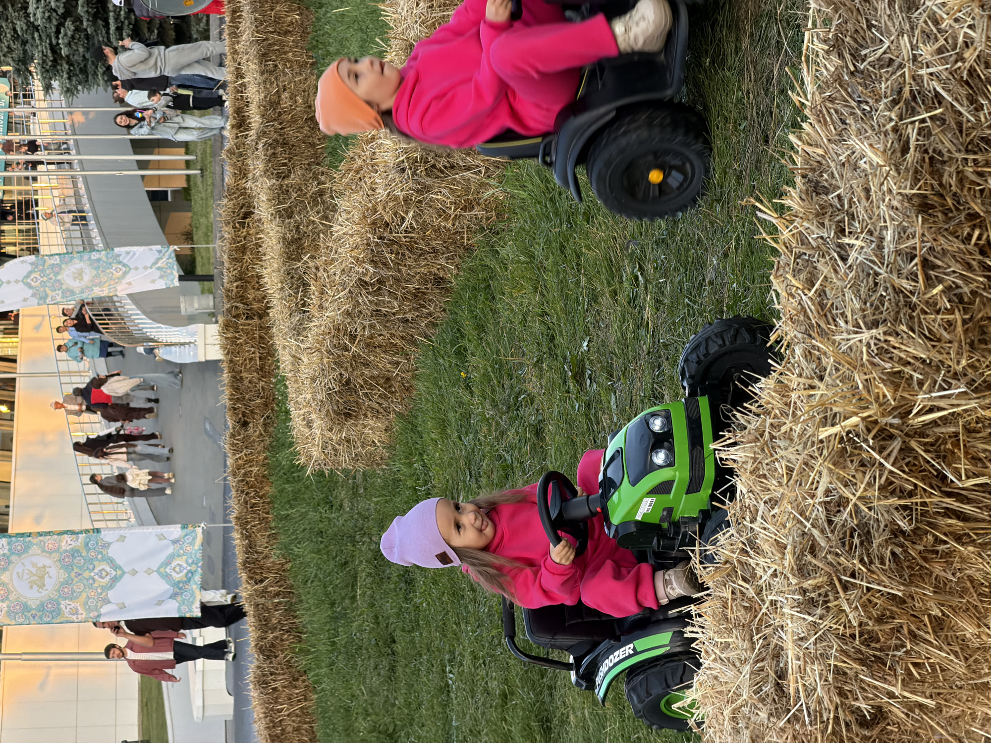Two young girls are riding on toy tractors in a park-like setting. The girl on the left is wearing a pink tracksuit and a purple beanie, and is sticking her tongue out. The girl on the right is wearing a pink tracksuit and an orange beanie. Both tractors are green and black. In the background, there are several people walking and standing around, and large bales of hay are scattered throughout the grassy area. There are also some decorative banners visible in the background. The lighting suggests it is either early morning or late afternoon.