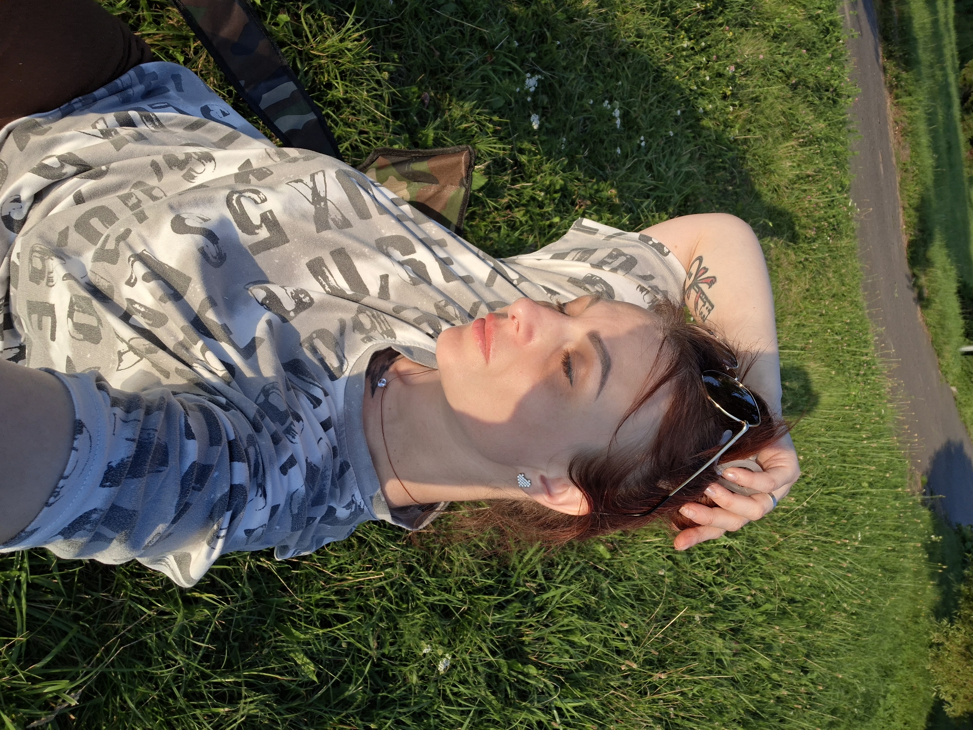 A woman is lying on her back in the grass, resting her head on her arm. She is wearing a grey t-shirt with a black and white letter print and sunglasses are resting on her head. Her eyes are closed, and her face is turned upwards towards the sun, which casts a warm glow on her skin. The grass is green and lush, with a few small white flowers scattered throughout. A paved path is visible to the right of the frame, with shadows of trees falling across it. The scene suggests a peaceful, relaxed moment outdoors, possibly in the late afternoon or early evening due to the quality of the light. There is no visible text in the image.