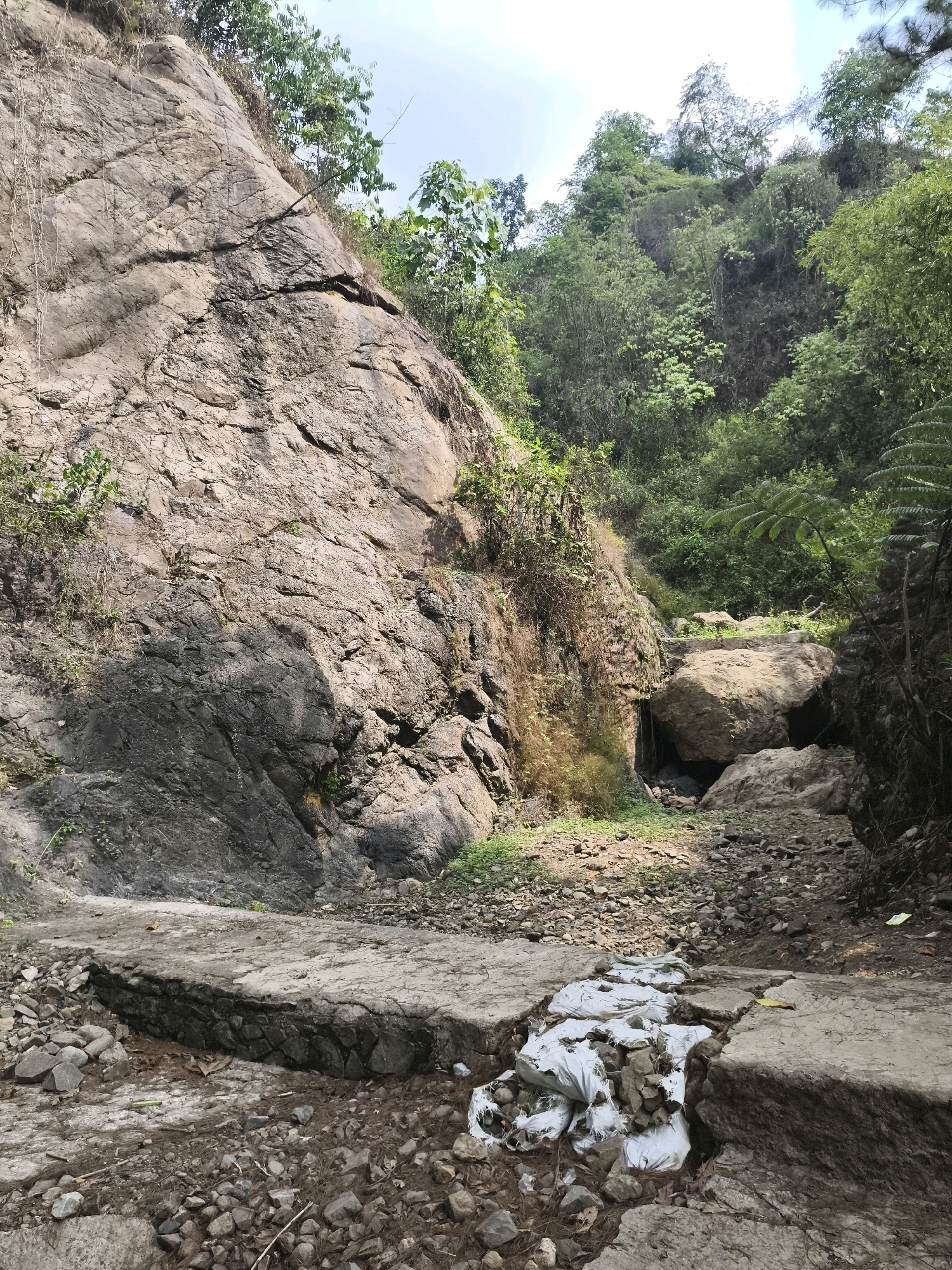 The image depicts a natural landscape in Babakan Madang, Indonesia, characterized by a steep, rocky cliff face on the left. Vegetation, including trees and vines, grows on the cliff and the surrounding hills. A dry, rocky streambed runs through the center of the scene, with several large boulders. In the foreground, a man-made concrete structure, possibly a pathway or retaining wall, has partially collapsed. Several white bags filled with debris, likely rocks and soil, are placed on the broken concrete, suggesting some form of repair or clean-up effort. The overall scene is one of a rugged, natural environment with signs of human intervention.