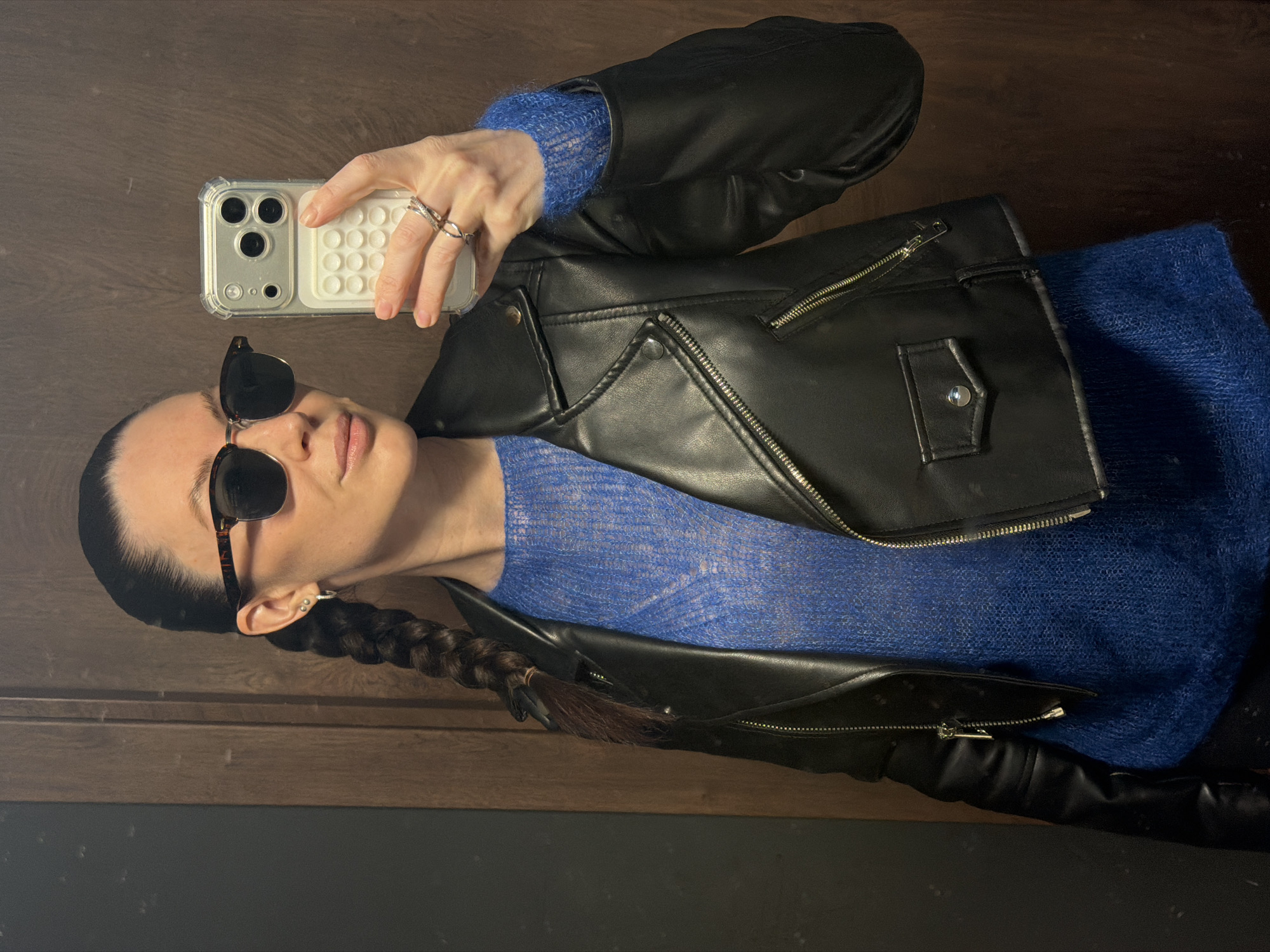 A person with dark hair braided and wearing sunglasses, a blue fuzzy sweater, and a black leather jacket is taking a selfie with a white smartphone. The background appears to be a wooden wall over a dark-colored wall. There is no information to identify the location as Perm, Russia.