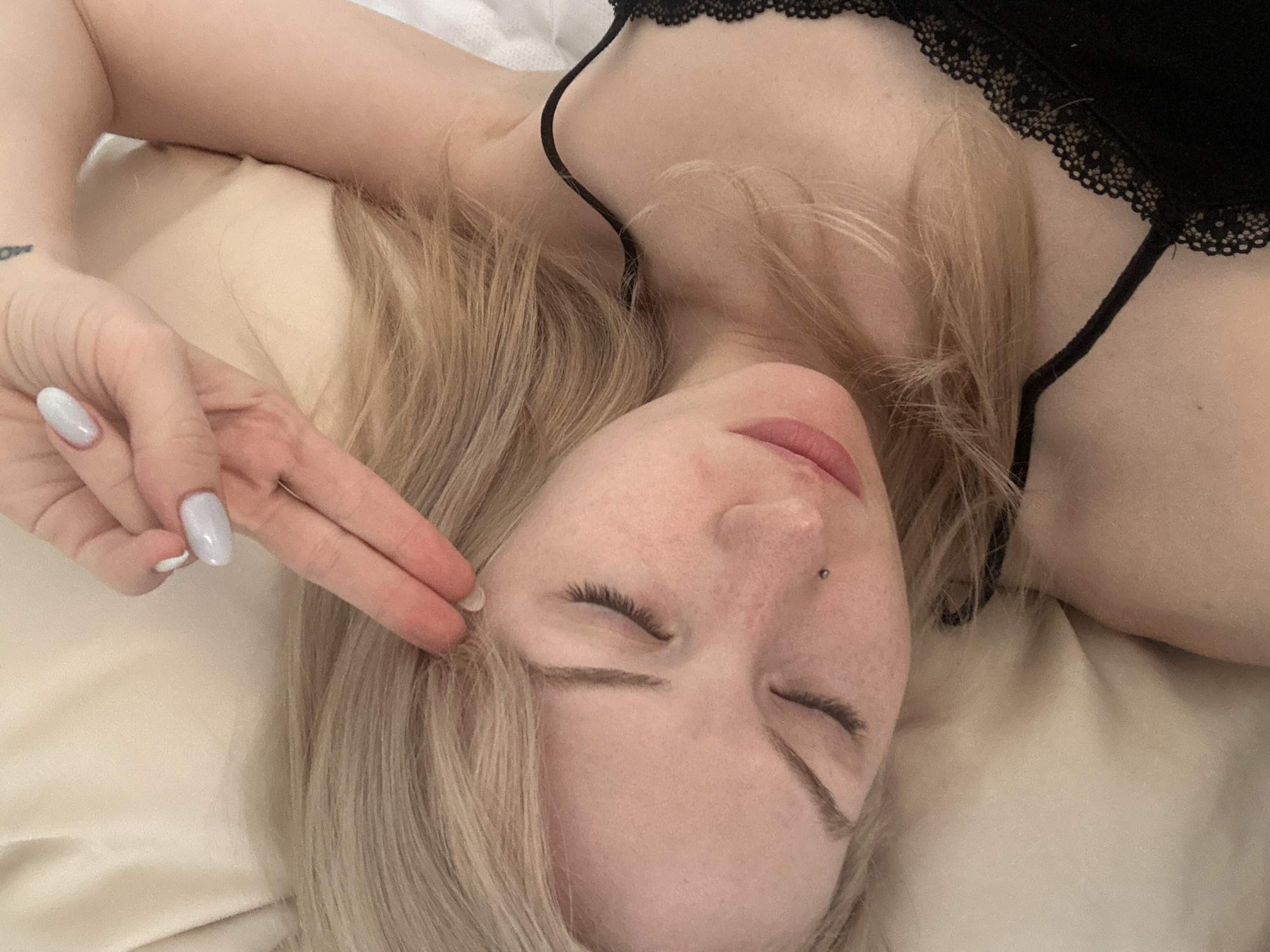 An overhead, medium shot shows a person with blond hair lying on their side on a light-colored pillow and bed. The person is wearing a black top with lace trim. Their eyes are closed, and their right hand is raised to their forehead with two fingers extended. The person has light skin with freckles and a nose ring. The background is out of focus, showing a cream-colored pillow and white sheets. No specific location in Tbilisi, Georgia can be determined from the image.