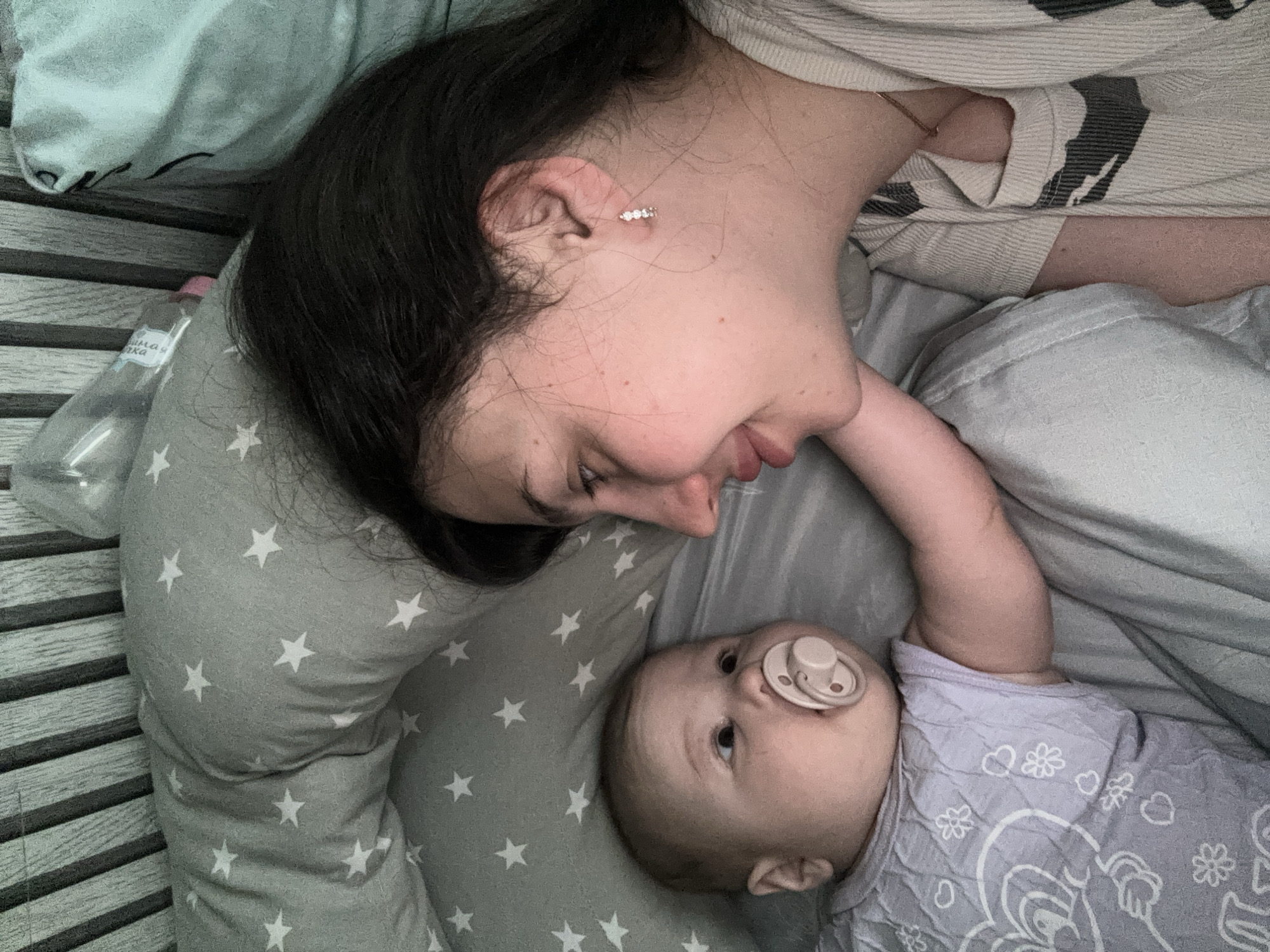 A mother and baby are lying in a crib. The mother has dark hair, fair skin, and is wearing an earring. She is looking down at the baby with a smile. The baby has fair skin, a pacifier in its mouth, and is looking up at the mother with its arm raised. They are lying on a gray sheet with white stars. A water bottle is next to them. The crib has gray wooden slats. The location is Марковский, Russia.