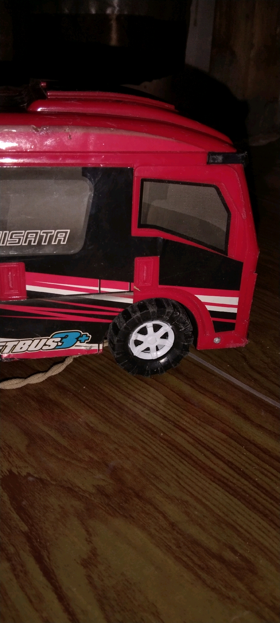 The image shows a close-up, eye-level, vertical shot of a red toy bus on a wooden floor. The bus is red with black accents and white stripes. Text on the side of the bus reads "WISATA" and "TBUS3+". A single wheel with a white rim and black tire is visible on the right side of the bus. The background is dark and out of focus, with some light streaks suggesting a wall or shelving. A thin, tan cord extends from the front of the bus. The scene appears to be indoors, on a wooden floor with a wood grain pattern. The location is identified as Unknown, Indonesia.