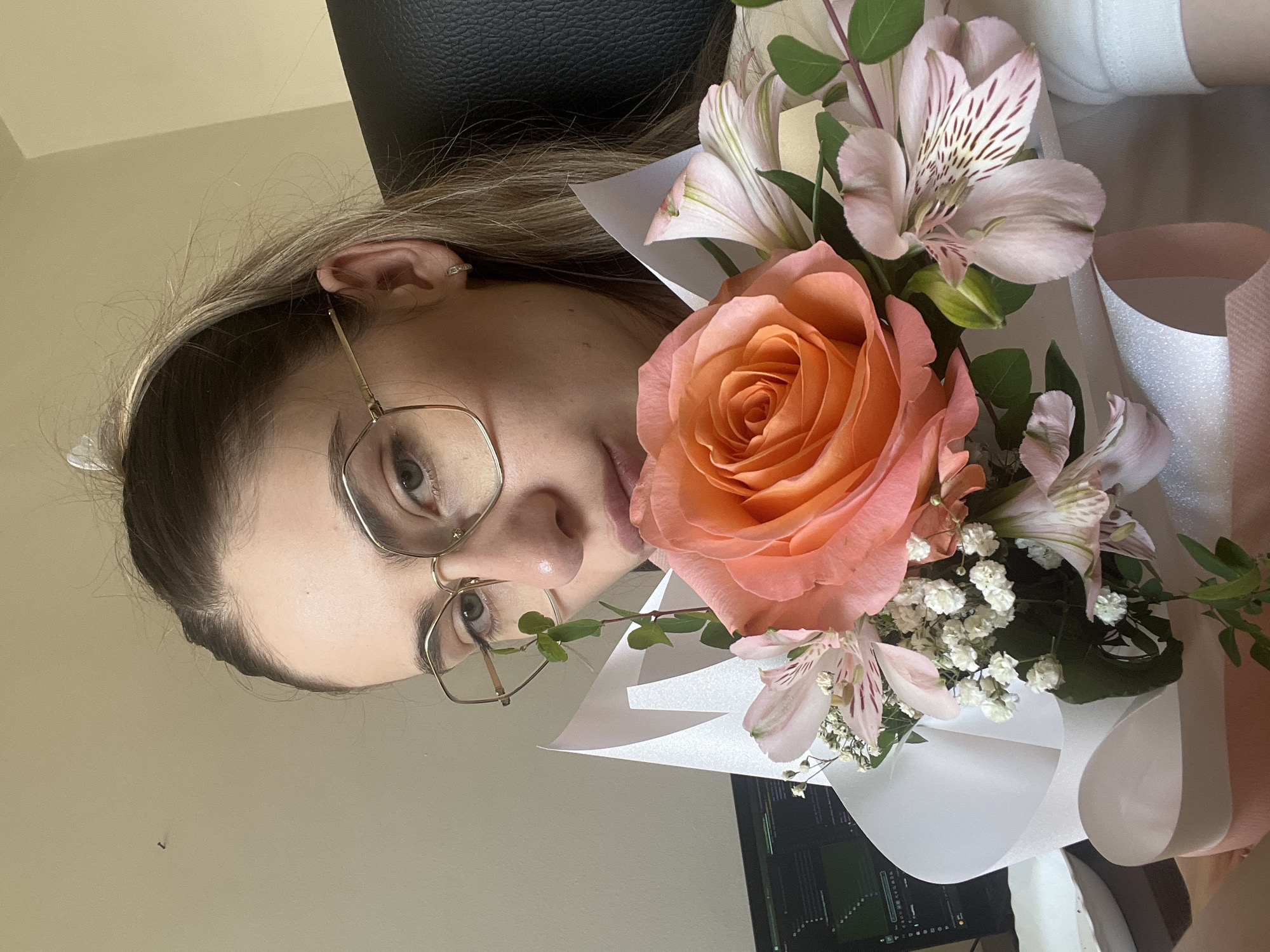 A young woman wearing glasses and a white shirt holds a bouquet of peach roses and pink alstroemeria flowers. She is seated in what appears to be an office or home office setting, with a computer monitor visible in the background. The lighting suggests an indoor environment.