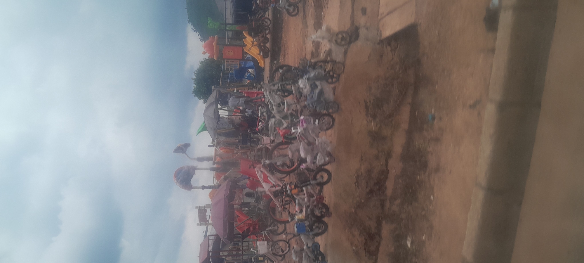 The image depicts an outdoor marketplace in Kugbo, Nigeria, characterized by a cloudy sky and a slightly elevated terrain. The foreground features a dirt road with a raised concrete curb. In the middle ground, a dense collection of motorcycles, bicycles, and what appear to be playground equipment are displayed. Some of the motorcycles and bicycles are covered in plastic. Several small stalls with umbrellas and canopies are set up, providing shade. A few individuals are visible, some standing and others appearing to be seated, suggesting commercial activity. The background reveals more structures and greenery, with hills visible in the distance. The overall impression is one of a busy, informal market selling various goods.