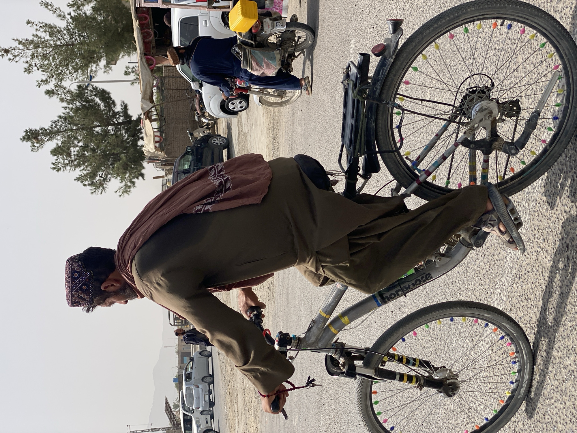 In Kandahar, Afghanistan, a man wearing a traditional embroidered cap and a brown tunic rides a bicycle. He is seen from behind, his face turned to the right. The bicycle has colorful beads attached to its spokes. In the background, several cars and motorcycles are visible on a dusty road. The environment appears to be an urban setting with low-lying buildings and sparse vegetation. The lighting suggests it is daytime, likely a sunny day with a clear sky.