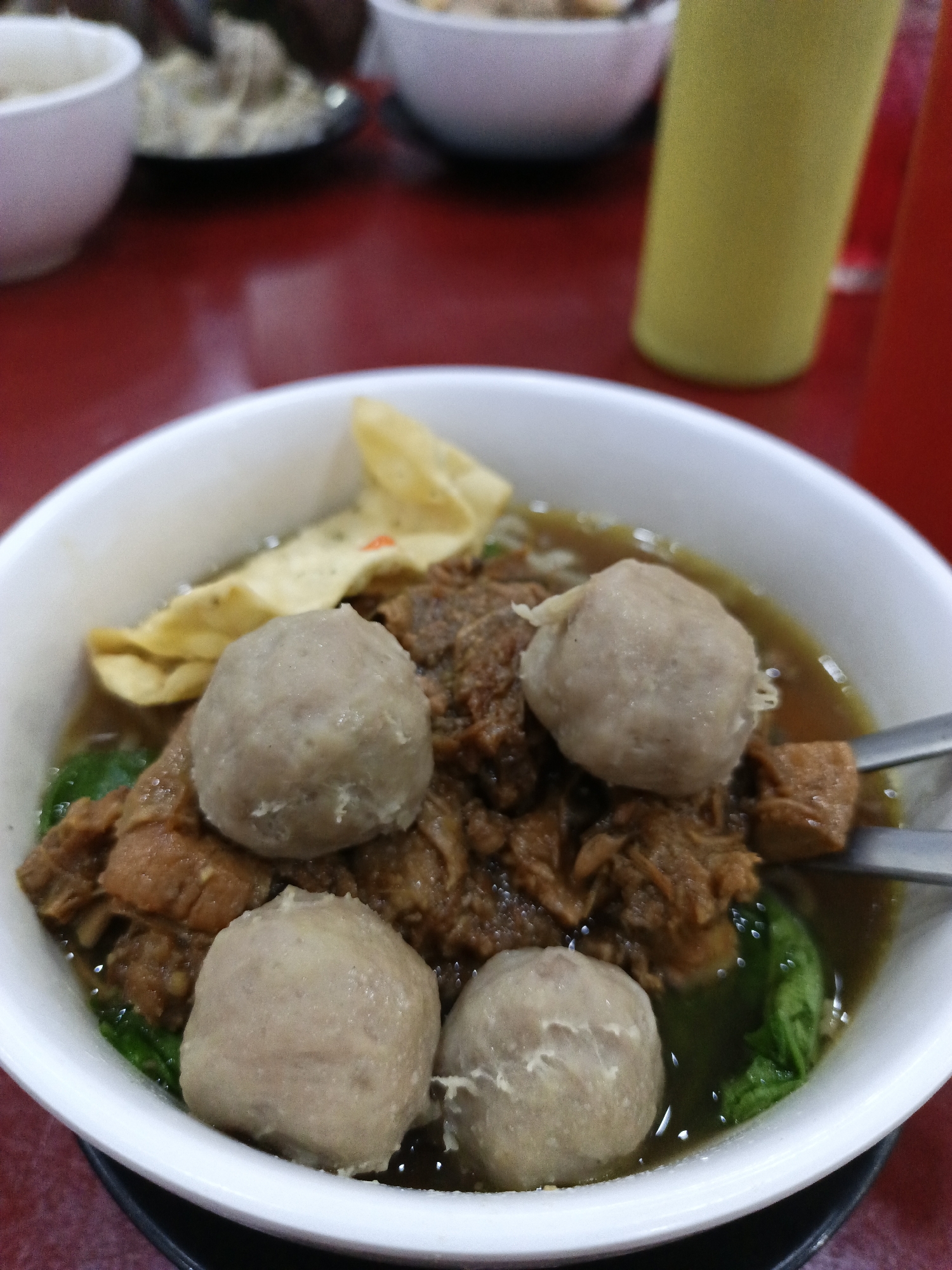 A close-up shot shows a white bowl filled with a soup dish. The bowl is filled with four light-brown meatballs, dark-brown braised meat, green leafy vegetables, and a fried wonton wrapper. The soup is light-brown. A metal spoon rests in the bowl.
The bowl sits on a black saucer. The background, out of focus, includes other bowls, a yellow bottle, and a red table.
The image is likely taken in Lhokseumawe, Indonesia.