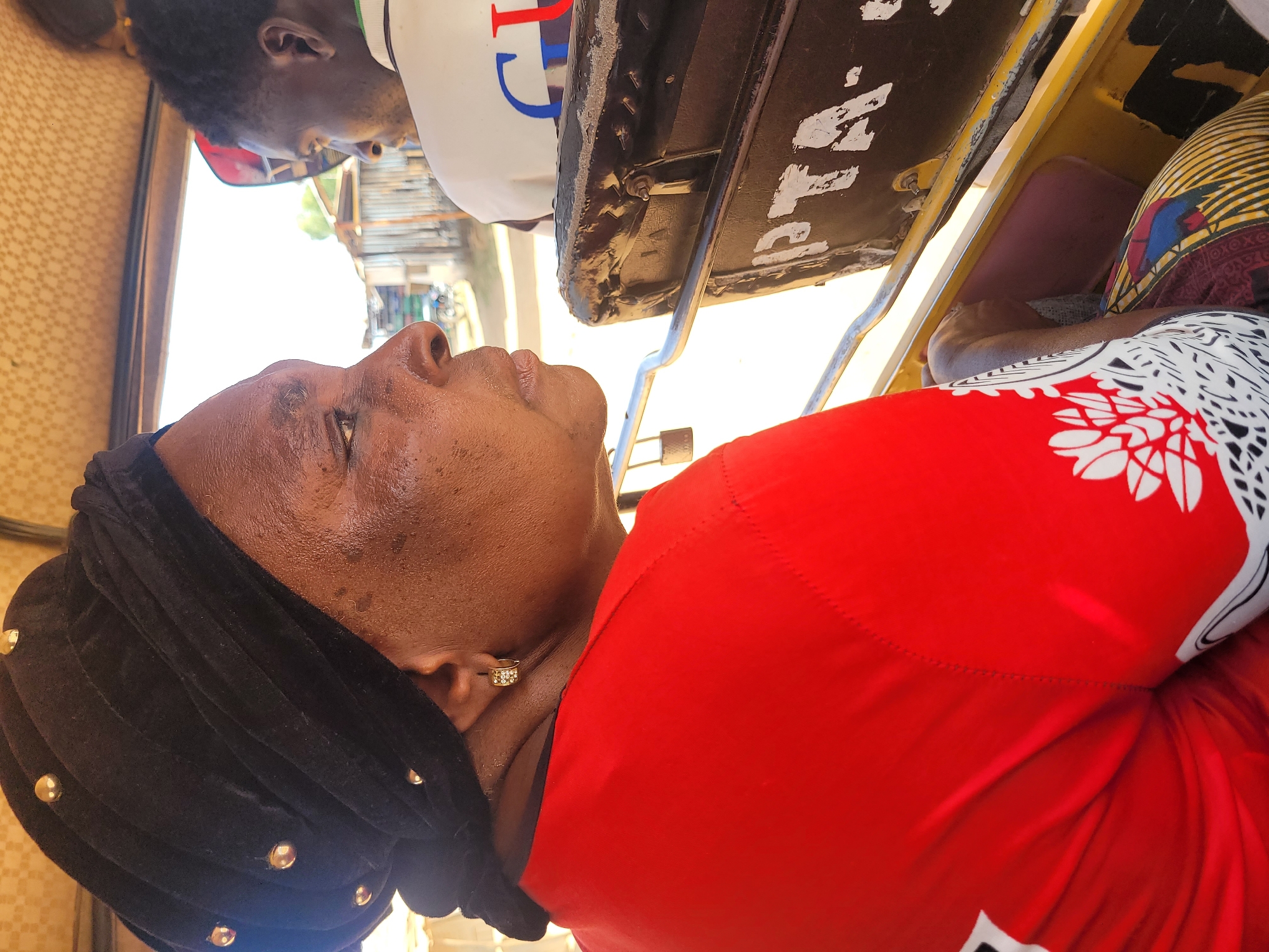 An eye-level, medium shot shows a woman riding in a yellow tricycle taxi in Jos, Nigeria. The woman is older with brown skin and is wearing a red shirt with a white floral design, along with a black velvet head covering adorned with gold studs. She is also wearing gold earrings. She is looking up. Seated next to her is a younger black man wearing a white T-shirt and red sunglasses. The taxi has a black seatback. The background shows a street scene.