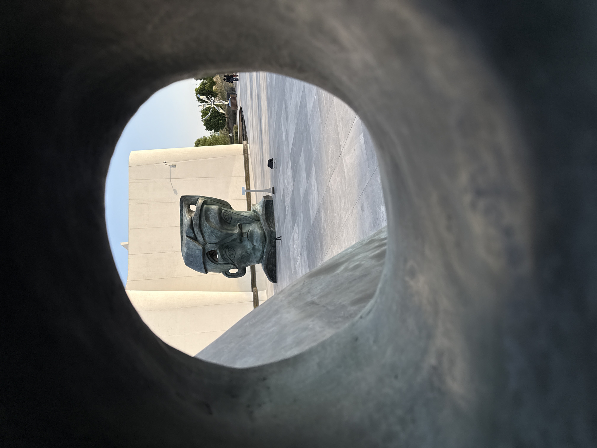 From a low angle, a bronze head sculpture is viewed through a circular opening in another sculpture. The head sculpture features a stylized human face with eye sockets and mouth. The background reveals a building with a light-colored exterior, a tiled walkway, and a distant sculpture of three intertwined elements, likely in San Bernardino Tlaxcalancingo, Mexico.