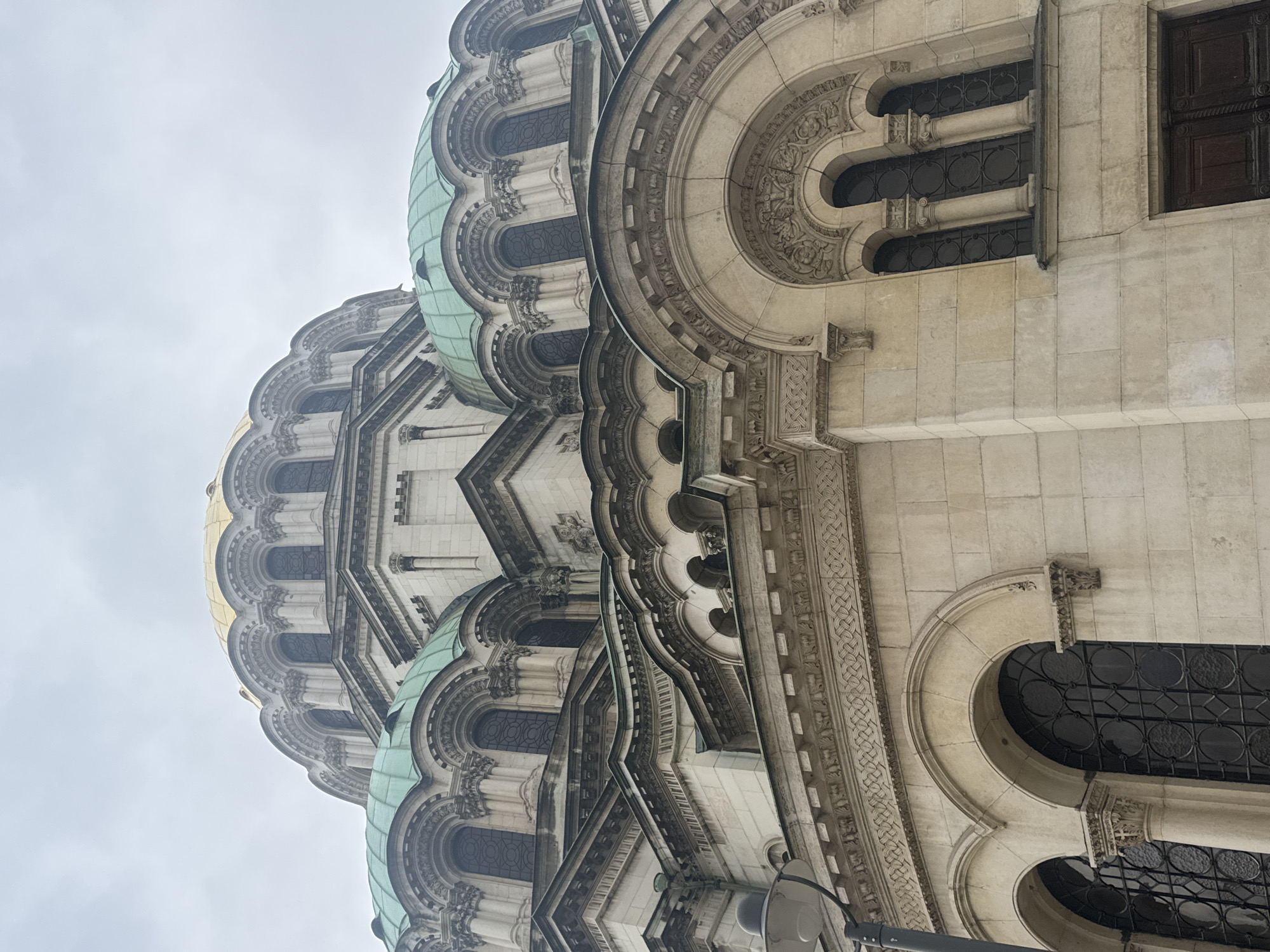 The image captures a low-angle view of the Alexander Nevsky Cathedral in Sofia, Bulgaria. The focus is on the architectural details of the cathedral, including its domes, windows, and ornate stone carvings. The domes are partially covered in green and gold. The sky is overcast, and the building is rotated, giving a distorted perspective.