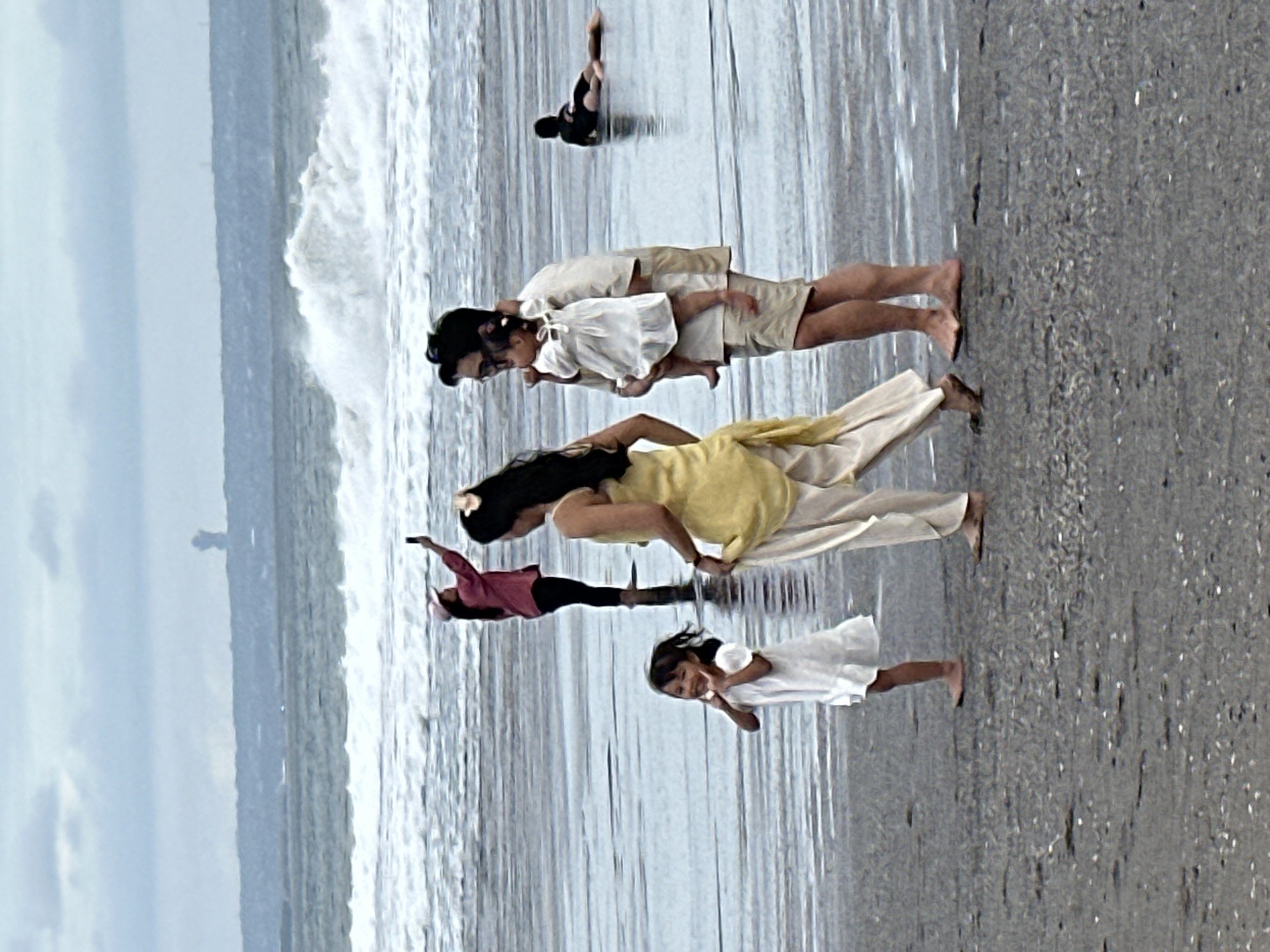 This image depicts a family on a beach in North Kuta, Indonesia. The sky is overcast, and the ocean has large waves crashing ashore. In the foreground, a young girl in a white dress stands on the wet sand, smiling and posing with her hands near her face. Behind her, a woman in a yellow top and white pants walks away from the camera, holding onto the hem of her pants. To the right of the woman, a man is holding a baby dressed in white. Further in the background, a person in a red shirt and a white hat is taking a photo with their phone, facing away from the viewer. Another person is lying on the wet sand in the distance. The beach is wet and appears to be composed of dark sand with small shells.