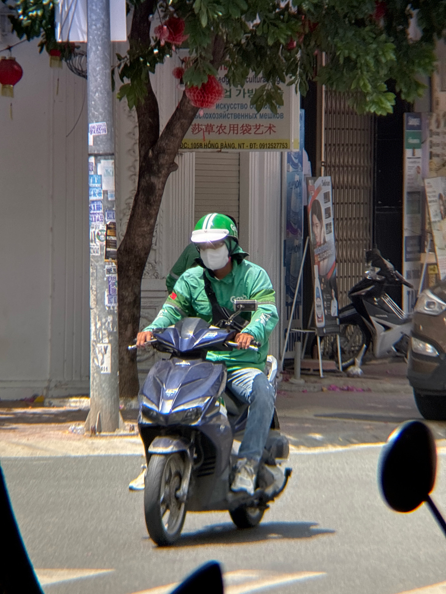 A person wearing a green jacket, white mask, and green helmet rides a dark blue scooter down a street in Nha Trang, Vietnam. The scooter is in the foreground, and the rider's legs and feet are visible. The rider is looking ahead, and their hands are on the handlebars. In the background, there are trees, buildings, and other scooters. There are also some signs with Vietnamese writing on them. The scene appears to be a sunny day, and the street is paved with asphalt. The image was taken from a low angle, possibly from another vehicle.