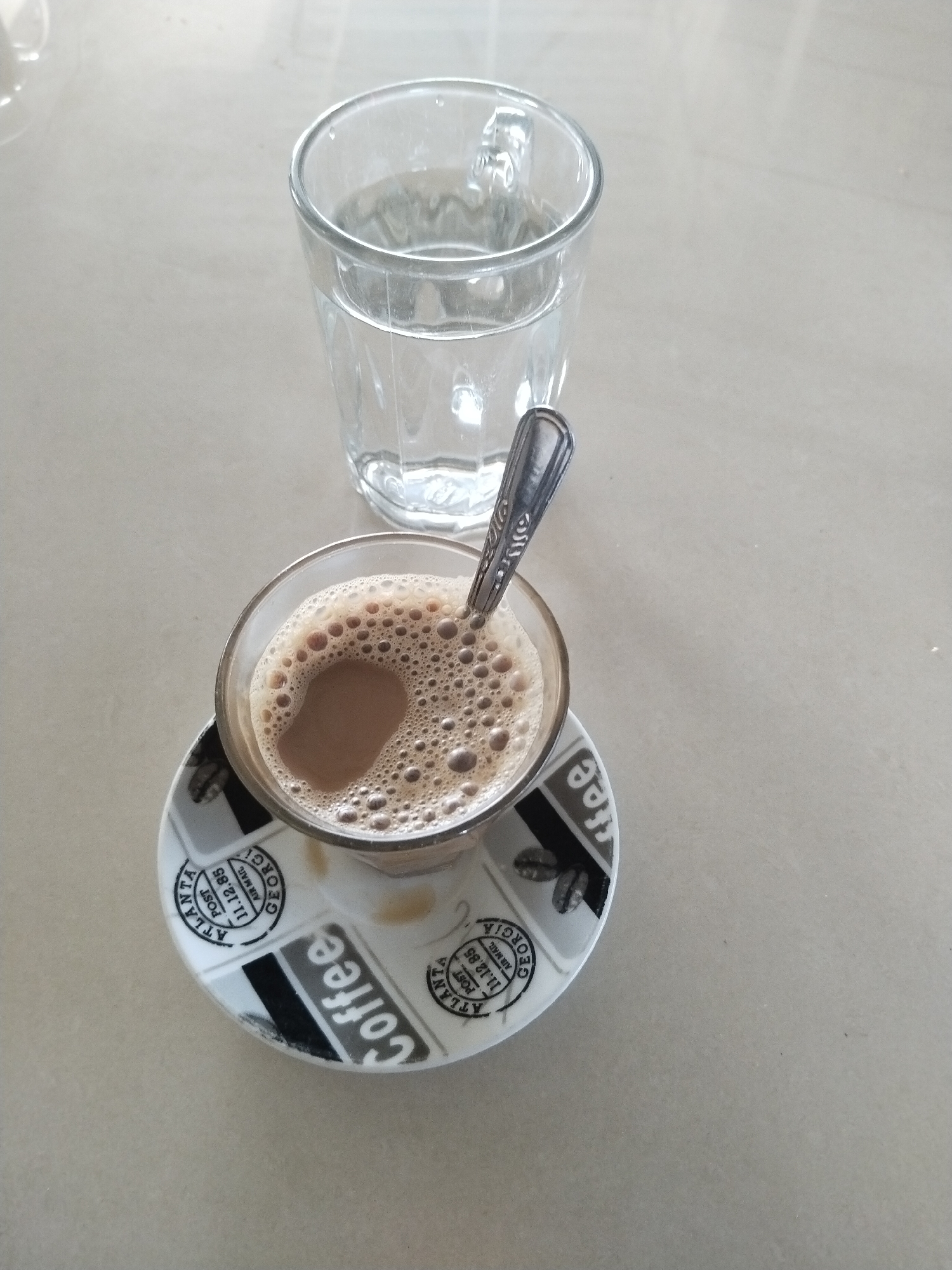 This image, taken in Matangkruet, Indonesia, displays a refreshing beverage setup from an overhead perspective. A light-colored, plain surface, likely a table or countertop, serves as the background.
In the lower center of the frame, a clear glass cup holds a light brown, frothy beverage, which appears to be coffee or a milky tea, topped with numerous small bubbles. A small, silver spoon with a decorative handle rests within the cup. This cup is placed on a round, white saucer. The saucer is adorned with black and grey graphics, including scattered coffee beans and text. Prominently visible text on the saucer includes the word "Coffee" in large, stylized letters. Additionally, there are circular stamp-like designs featuring text such as "ATLANTA POST," "GEORGIA," "AIR MAIL," and a date "11.12.85."
Behind and slightly to the left of the coffee cup, a clear glass is filled with plain water, reflecting the ambient light. The overall scene suggests a moment of casual refreshment.