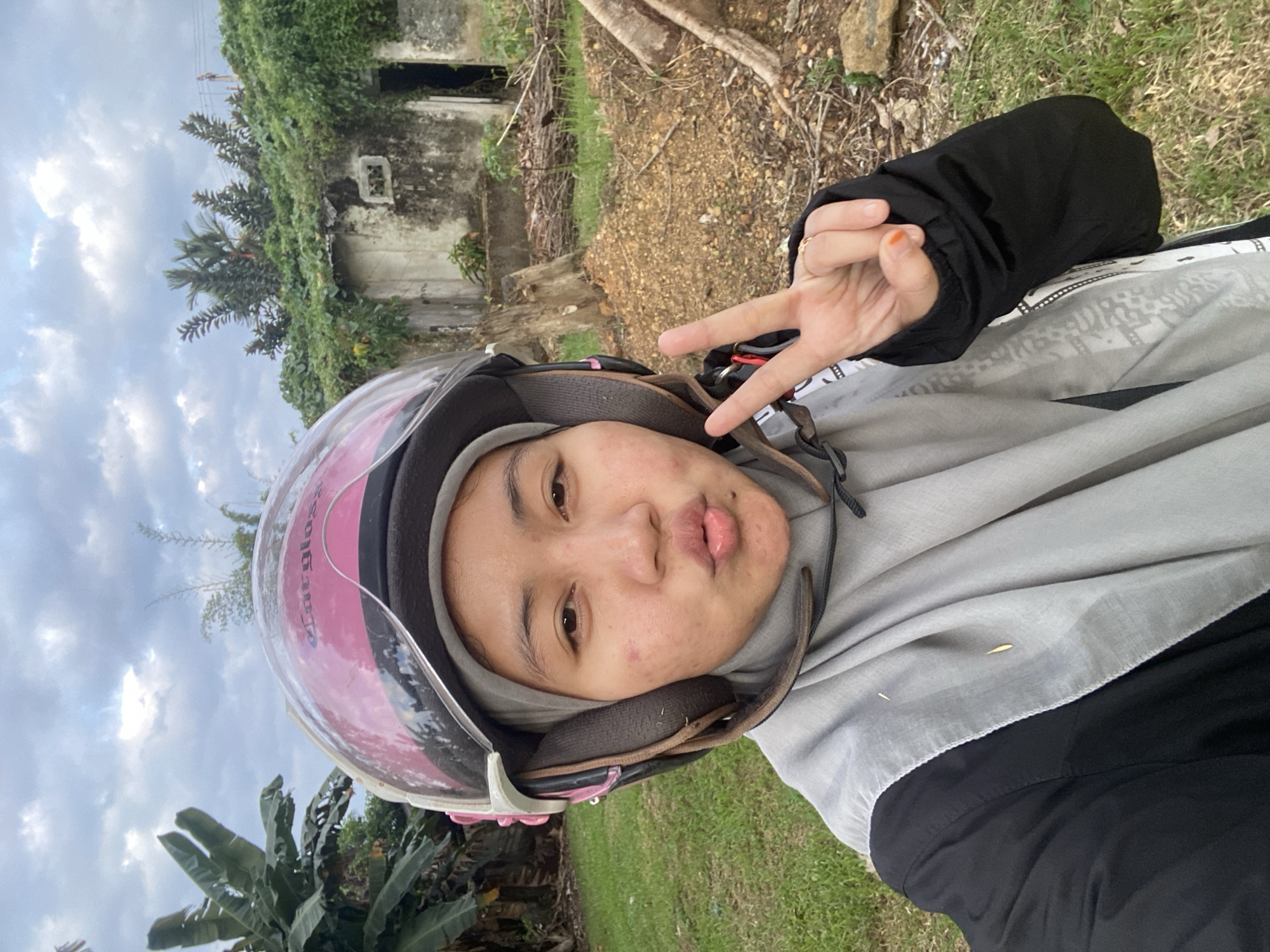 A young woman, wearing a grey hijab and a black jacket, is taking a selfie outdoors. She has a pink helmet on her head and is making a peace sign with her right hand. Her lips are pursed. Behind her, there is an overgrown, abandoned building with green foliage covering it. Banana trees are visible to the left of the frame, and the ground is covered in dry leaves and dirt. The sky is cloudy. This scene is consistent with the rural outskirts of Banjarbaru, Indonesia, where such abandoned structures and dense vegetation are common.