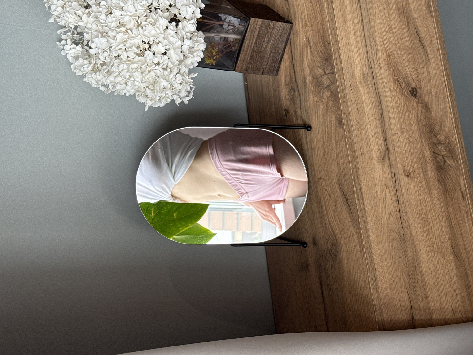 The image shows a top-down view of a tabletop setting. Located on the left side is a gray surface, likely a countertop. Adjacent to it is a wooden-patterned surface, possibly a cabinet or a table. A bouquet of white hydrangeas sits near a small, dark, decorative wooden box.
A round mirror on a stand reflects a torso wearing a white shirt and pink bra. A green leaf partially obscures the reflection. The reflection also shows a window and building behind it, suggesting the photograph was taken in an interior setting in a location with multiple residential buildings.
The precise location is not evident from the image, and no definitive details confirm that it is Chaikovsky, Russia. The reflection shows a window and buildings behind it, suggesting the photograph was taken in an interior setting.