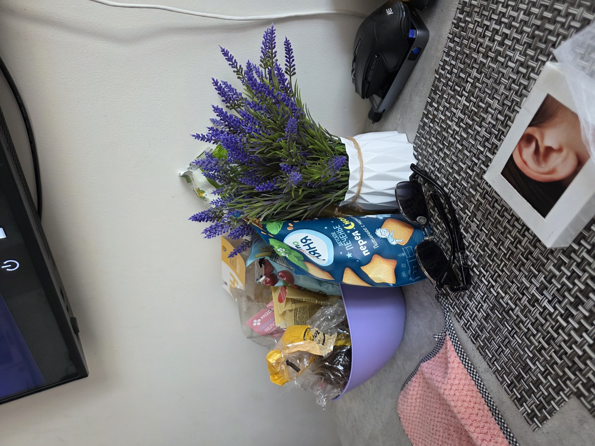A still life arrangement is depicted on a gray, textured surface, likely a table. In the background, a light-colored wall serves as a backdrop. Dominating the center of the scene is a light purple bowl filled with various snack packages, some transparent and others with colorful labels. To the right of the bowl, a white, geometric vase holds a vibrant bunch of artificial lavender.
In the foreground, a textured pink cloth with a black and white checkered trim lies on the left. Next to it, a pair of dark sunglasses rests on a dark gray woven placemat. To the right of the placemat, a black computer mouse sits on top of a gray device. Further to the right, a white box with an image of a human ear is partially visible.
The overall impression is a domestic setting, possibly a living room or kitchen counter, with items suggesting casual snacking and personal accessories. The scene does not provide any specific indicators of its location within Sochi, Russia.