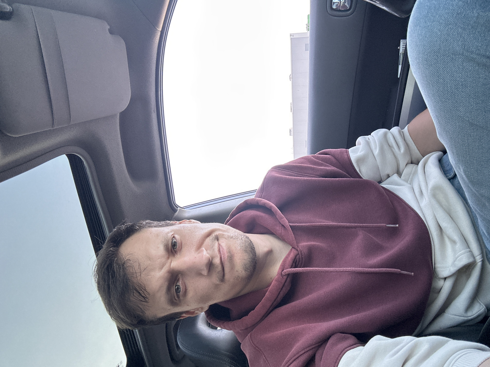 A man is sitting in the driver's seat of a car with a sunroof open. He is wearing a maroon and cream hooded sweatshirt and blue jeans. The man is looking directly at the camera with a neutral expression. The interior of the car is visible, including the dashboard, a portion of the steering wheel, and the car door. Outside the car window, a light-colored building and a clear sky are visible. There is no indication of the city of Kamianets-Podilskyi, Ukraine, in the image.