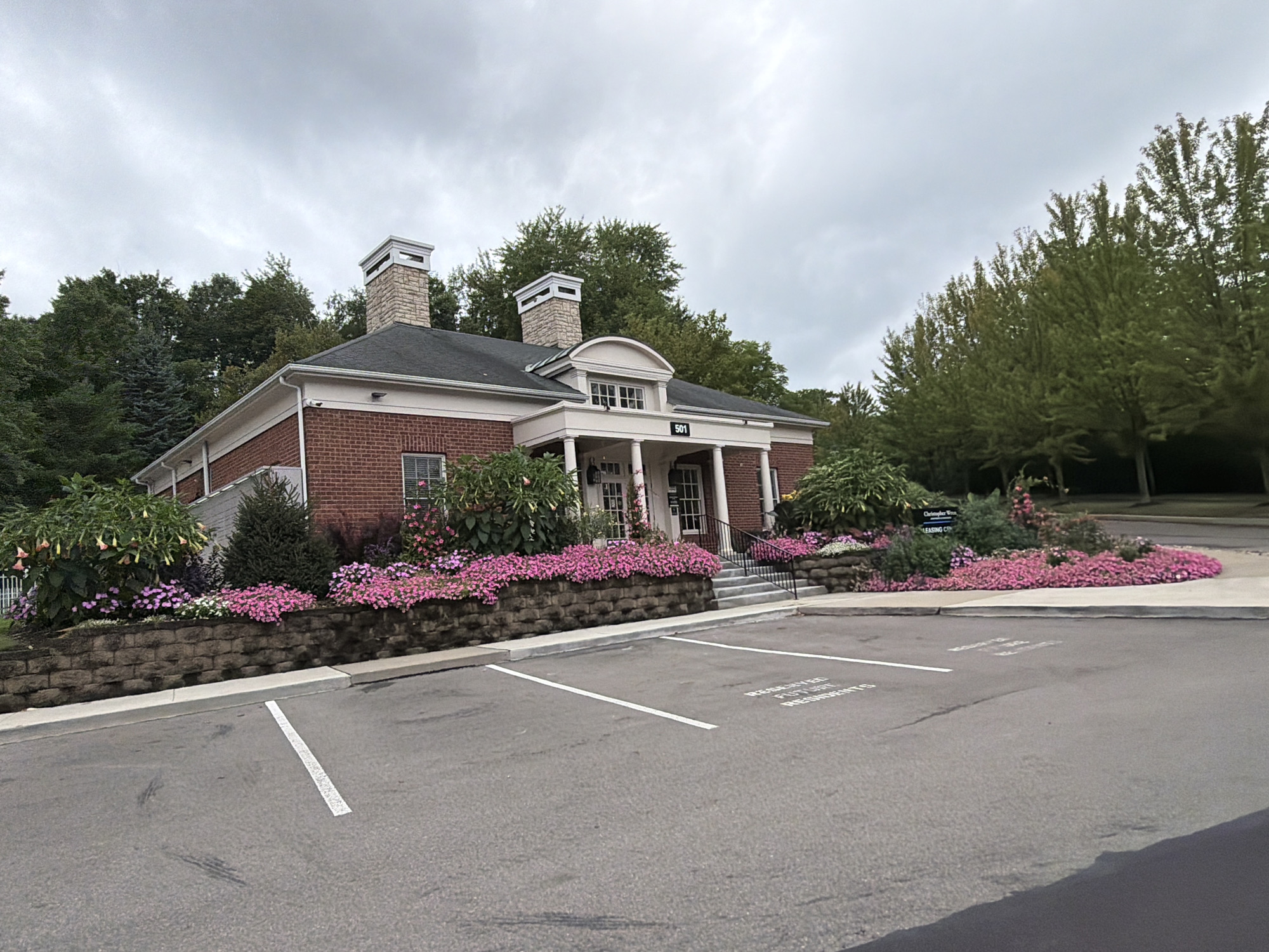 The image shows the exterior of a brick building with a white portico and a dark roof. The building has two chimneys and is surrounded by lush greenery and flowering plants, predominantly pink flowers lining a stone retaining wall. A parking lot with white lines is in the foreground, with one parking space marked "RESERVED FOR FUTURE RESIDENTS." The sky is overcast with gray clouds. The building appears to be the leasing office for a residential complex. Based on the sign visible in the image, the location is Christopher Woods, Leasing Center, in Wexford, United States of America.