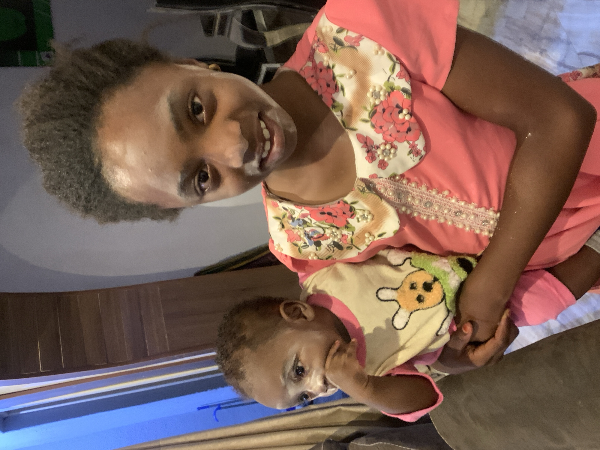 A woman with dark, short, curly hair smiles at the camera while holding a baby. The woman is wearing a pink dress with a floral collar and pearl accents. The baby is wearing a light yellow and pink onesie with a cartoon character on the front. The woman is holding the baby in her arms. The background is a room with a wooden cabinet and a window with blue trim. The image appears to be taken indoors. The location is Jos, Nigeria.