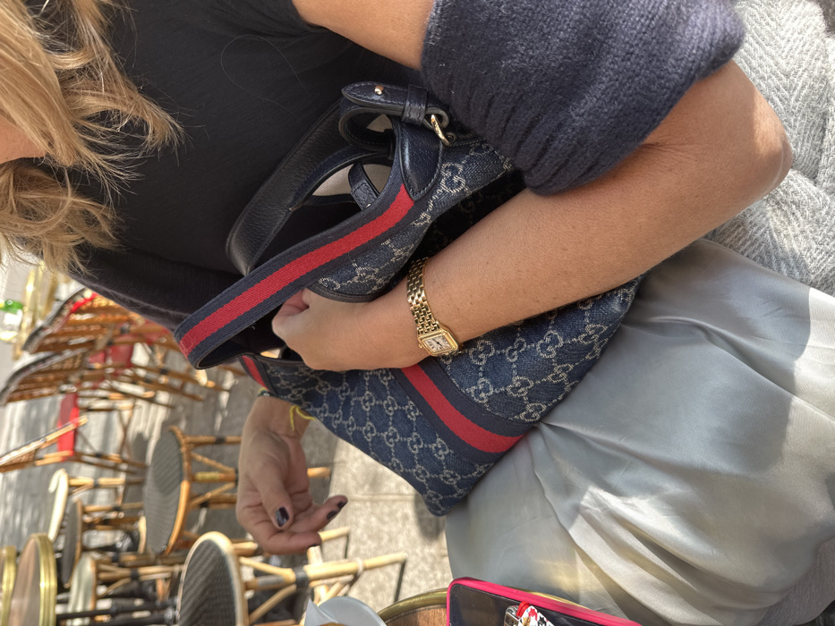 A seated individual is depicted from the chest down at an outdoor café in Paris, France. The subject has fair skin and visible blonde hair at the top of the frame. They wear a black top and a light-colored lower garment, with a textured grey blanket or throw draped over their lap.
The left hand, with dark-painted fingernails, rests on a blue denim-patterned shoulder bag featuring a repeating 'GG' monogram and a prominent red and dark blue striped web strap. The bag's opening is visible, with the hand partially positioned inside. The right wrist displays a gold-toned rectangular watch with a gold-toned link bracelet. To the lower left, a pink-rimmed object, consistent with a smartphone in a case, is visible on a table surface, accompanied by small white objects.
The background is blurred, showing typical outdoor café furniture, including multiple round tables with dark tops and wooden stools with woven cane seats. Additional chairs with red seats are partially visible further back. The scene is illuminated by strong natural sunlight, creating distinct shadows on surfaces.