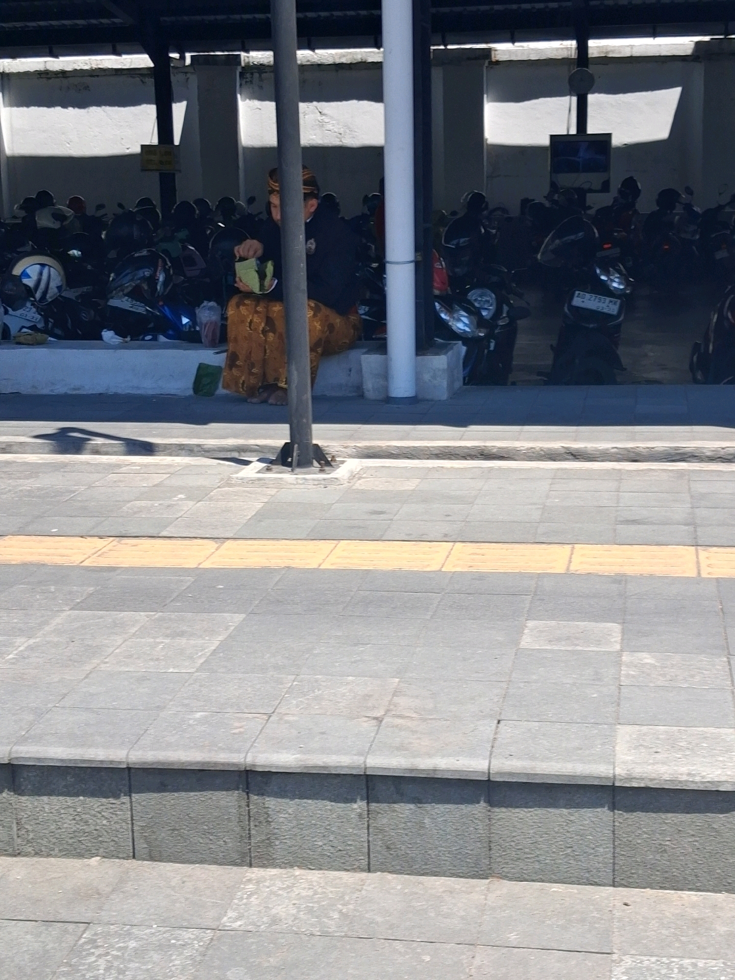 In Surakarta, Indonesia, a person wearing a traditional Javanese outfit, including a batik sarong and headwear, sits on a raised platform near a column. The individual is holding a small green object. The background reveals a parking area filled with motorcycles, suggesting a public space like a market or transport hub. The foreground displays a paved walkway with a distinctive yellow tactile paving strip, commonly found in pedestrian areas to aid visually impaired individuals. The scene is illuminated by sunlight, casting shadows on the ground.
