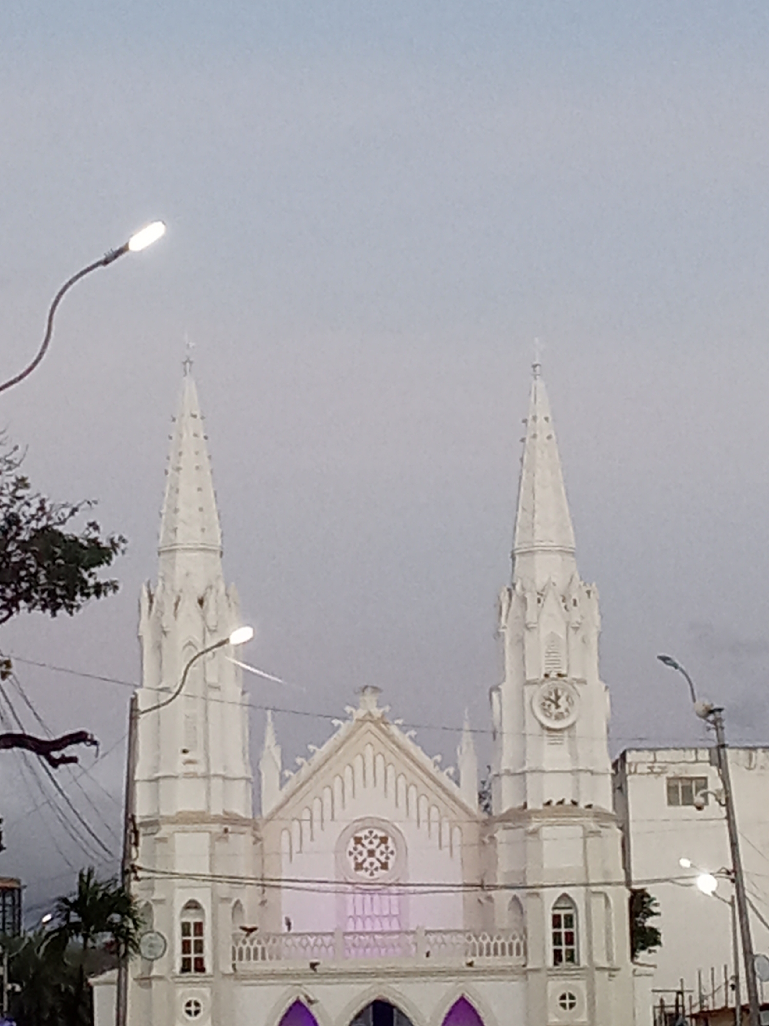 The image displays the upper section of the Church of San Juan Evangelista in Juan Griego, Venezuela, set against an overcast sky. The church is characterized by its white Gothic Revival architecture, featuring two prominent, pointed spires, both culminating in crosses. The right spire incorporates a clock face. The central facade includes a gabled section above a circular rose window, with multiple pointed arch windows, some illuminated with purple light. A white-railed balcony spans the lower visible portion of the central facade. In the foreground, two streetlights with bright white illumination are visible, one on the far left with a curved arm and another on the right attached to a utility pole. A network of dark utility wires crisscrosses the frame, and dark green foliage from trees or bushes is present on the left side. A plain, light-colored building is partially visible to the immediate right of the church. No individuals or discernible actions are depicted.
