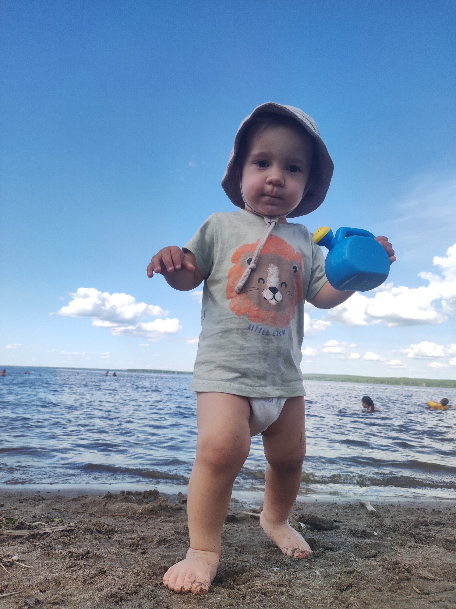 A young child, wearing a bucket hat and a t-shirt with a lion graphic, stands on a sandy beach. The child is holding a blue plastic watering can. The background shows a body of water with several people swimming or playing in it. The sky is blue with scattered white clouds, suggesting it is daytime with fair weather. The image appears to be taken at a lake or large river. The t-shirt has text that reads "LITTLE LION".