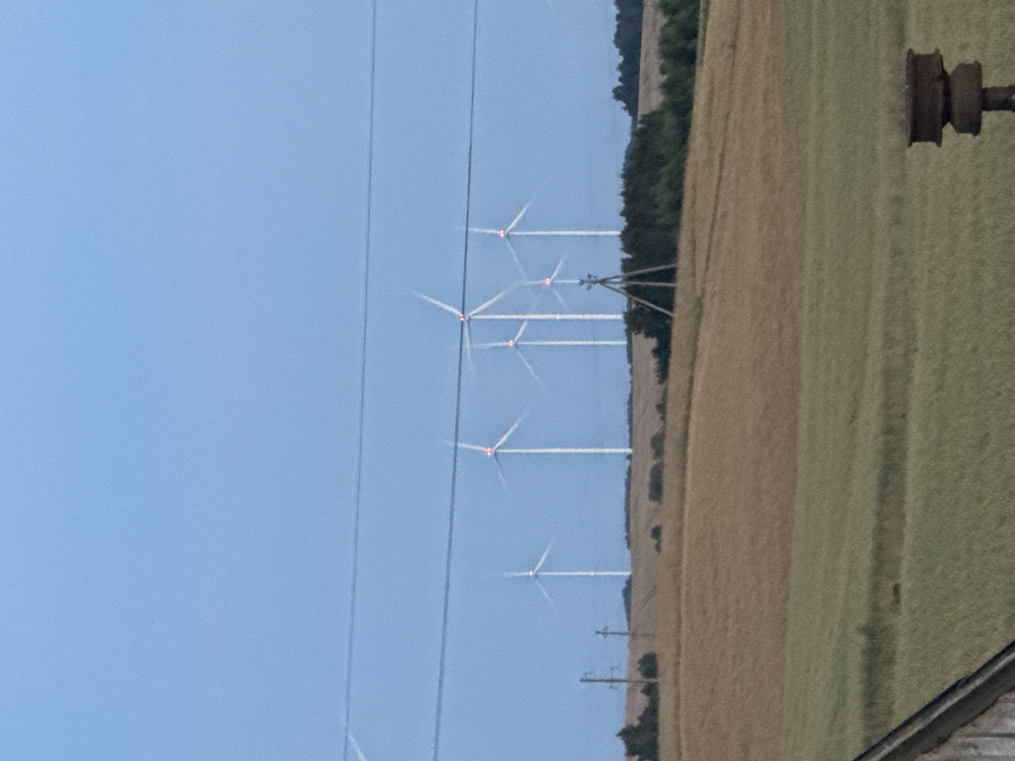 This is a photograph taken in Vikraičiai, Lithuania, depicting a landscape dominated by a wind farm. In the foreground, a power line stretches horizontally across the frame. Behind it, rolling fields of agricultural land are visible, with a section of what appears to be dry, harvested crops and another section of greener, growing crops. A few wooden utility poles are interspersed in the mid-ground. The background is filled with a line of wind turbines, their white towers rising against a clear, pale blue sky. The blades of several turbines are in motion, and some have red warning lights illuminated. The overall scene suggests an open, rural environment.