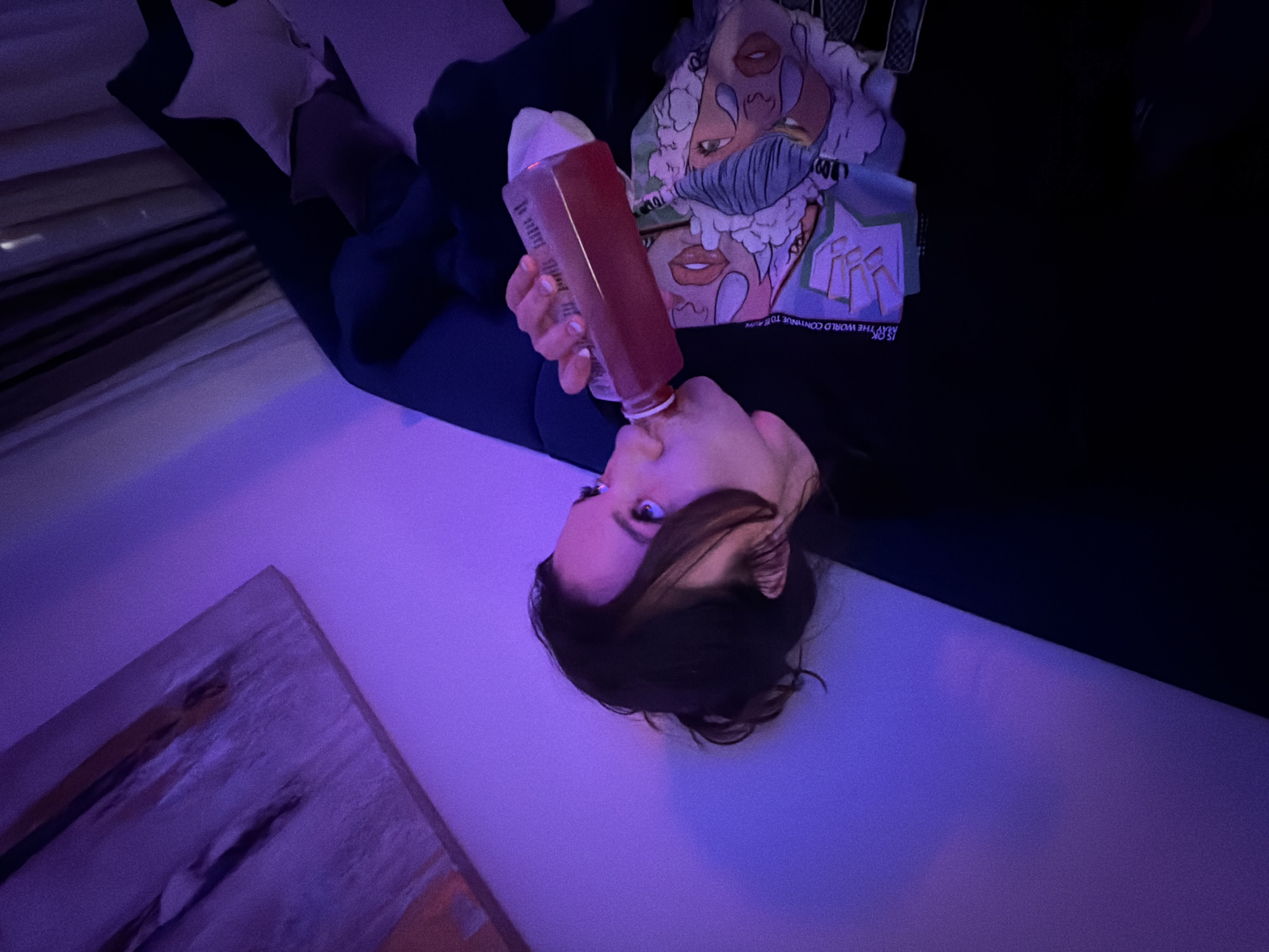 A woman is reclined, drinking from a red bottle. She is wearing a black top with a graphic print on the front. The scene has a purple hue, with a piece of artwork visible in the corner. It's impossible to determine the precise location within Belgrade, Serbia, from this image alone.