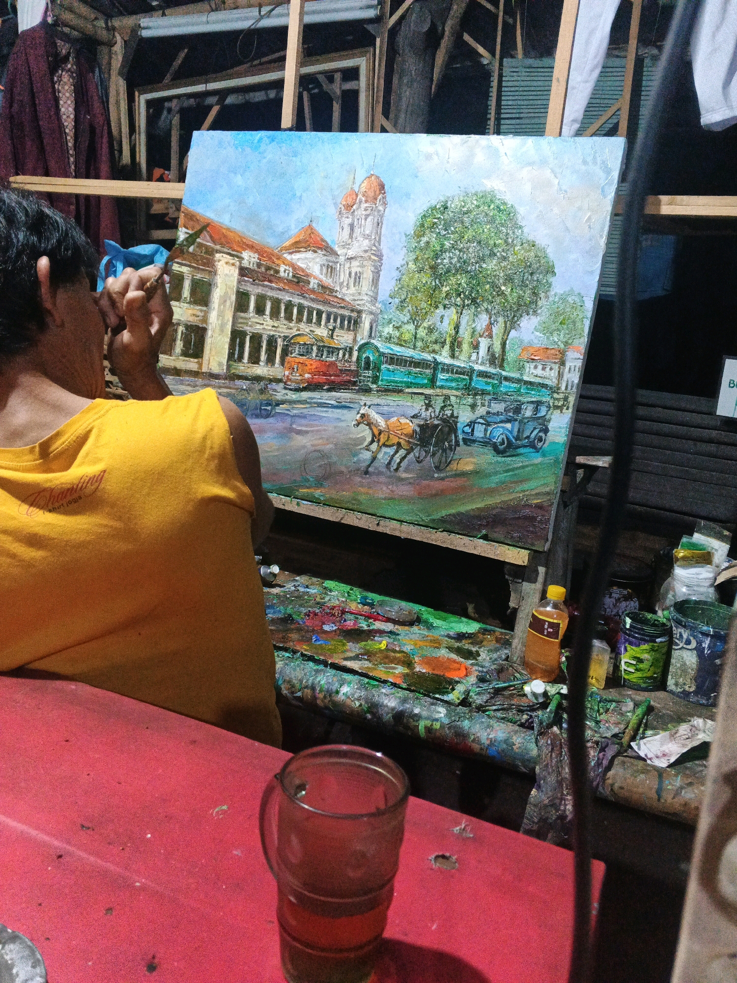 The image shows an artist in a dimly lit studio, likely in Ungaran, Indonesia, working on a vibrant oil painting. The painting depicts a street scene with a large colonial-style building with domed towers, a lush green tree, a horse-drawn carriage, an early automobile, and what appear to be trams. The artist, seen from behind, wears a yellow t-shirt and is holding a brush. The foreground features a red surface, possibly a table, with a glass of amber liquid and various art supplies, including a palette covered in colorful paints, jars of paint, and brushes. The background of the studio is cluttered with art materials, fabric, and wooden structures. The overall atmosphere suggests an active, creative workspace.