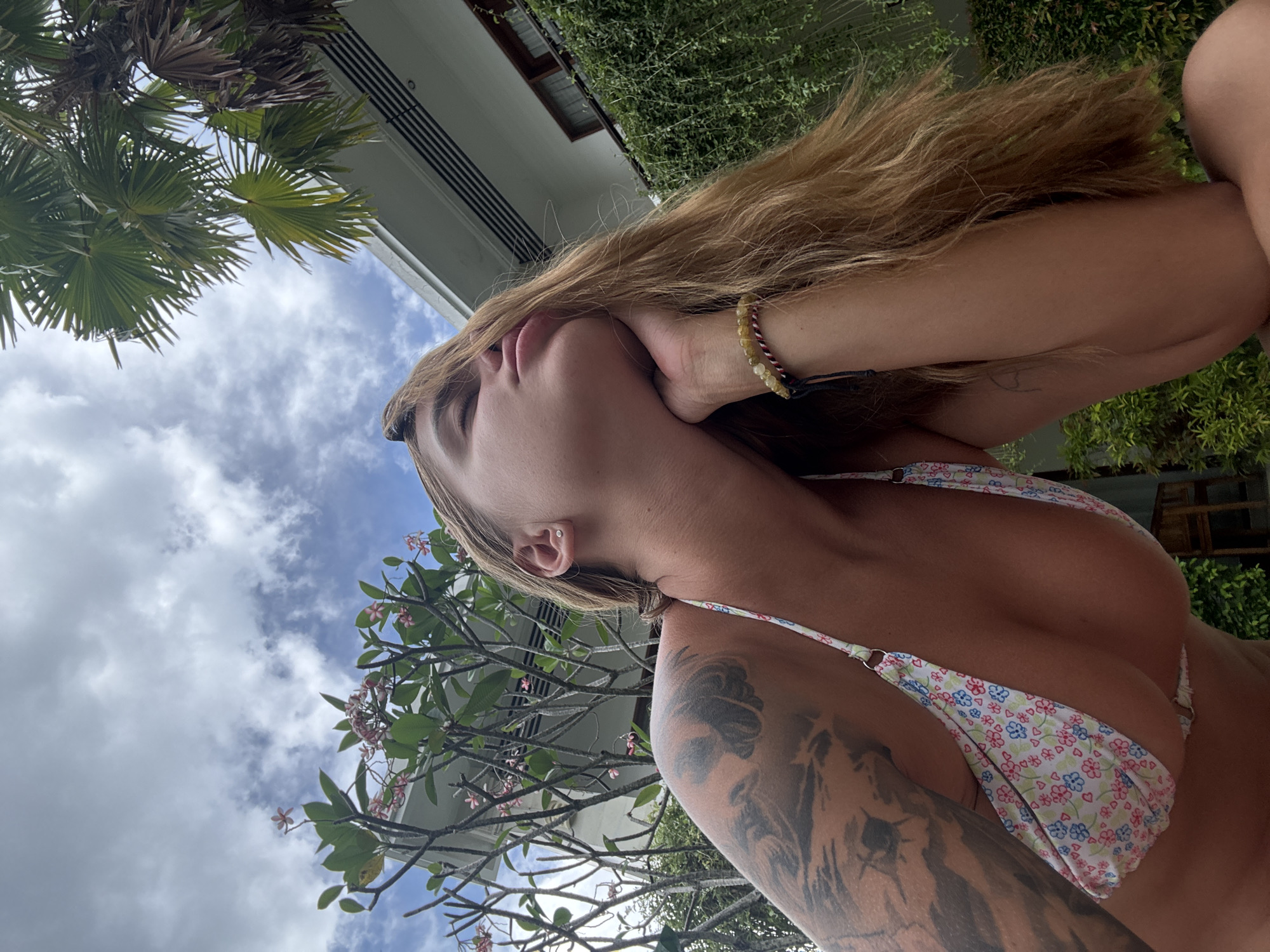 This image captures a woman in an outdoor, tropical setting, likely in North Kuta, Indonesia. The photo is taken from a low angle, looking upwards towards the subject and the sky. The woman has long, light brown hair, which flows around her head. She is wearing a white bikini top adorned with a delicate pattern of small red, blue, and green flowers and hearts. A prominent tattoo, appearing to depict a large feline like a tiger or lion, covers a significant portion of her left upper arm and shoulder. She has a small, silver stud earring in her left ear and a beaded bracelet on her right wrist.
The woman's head is tilted back, and her right hand is gently placed near her jawline, suggesting a posed shot. Her expression appears serene or relaxed. The background features a partly cloudy blue sky, indicating daytime. Lush green foliage, including large palm fronds and smaller leafy climbing plants, frames the upper and right sides of the image. A frangipani tree with delicate pink and white blossoms is also visible. Portions of a modern building with white walls and horizontal architectural details are present, hinting at a resort or contemporary structure. No text is visible in the image.