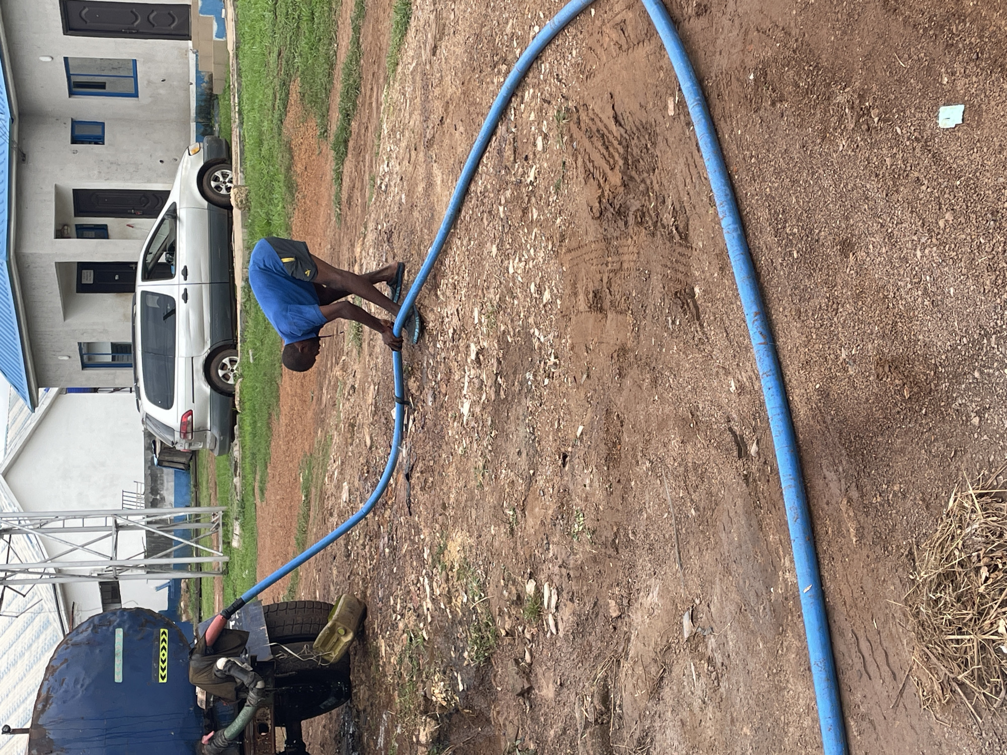 A person is seen bending over while holding a blue hose in an outdoor scene. The person is wearing a blue shirt and dark shorts. The setting appears to be a dirt ground with some patchy grass and scattered small stones. A white vehicle and a building with blue trim are visible in the background. There is also a large dark blue tank on a trailer with tires, suggesting it might be a water truck. The hose is connected to the tank and extends towards the person. The overall impression is that the person is operating or managing the hose, possibly for filling or emptying the tank. The lighting suggests it is daytime. There are no discernible emotions visible. There is some text on the side of the tank, which appears to be yellow and black hazard symbols.
