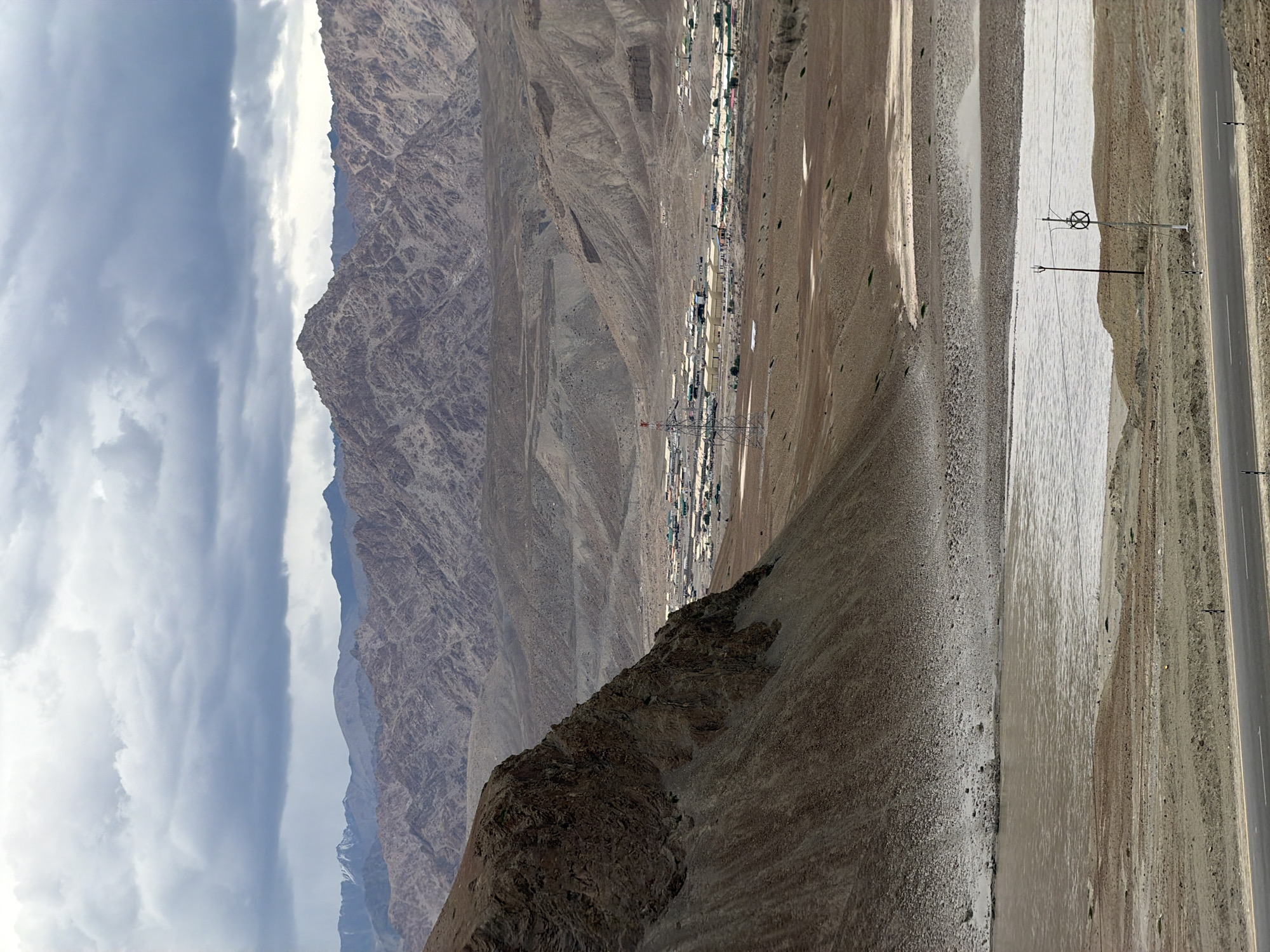 The image shows a wide, panoramic view of a mountainous landscape near Chumkar, India. In the foreground, a paved road runs horizontally across the bottom of the frame. To the left of the road, a river flows, its water appearing muddy brown. Beyond the river, the terrain is arid and rocky, with scattered patches of dry vegetation. In the mid-ground, a settlement is visible, characterized by numerous low-lying buildings with colorful roofs. A tall, red and white transmission tower stands to the left of the settlement, connected to power lines. The background is dominated by rugged, eroded mountains in varying shades of brown and gray, under a cloudy, overcast sky. The overall impression is one of a remote, high-altitude environment with a small human settlement nestled within a vast natural landscape.