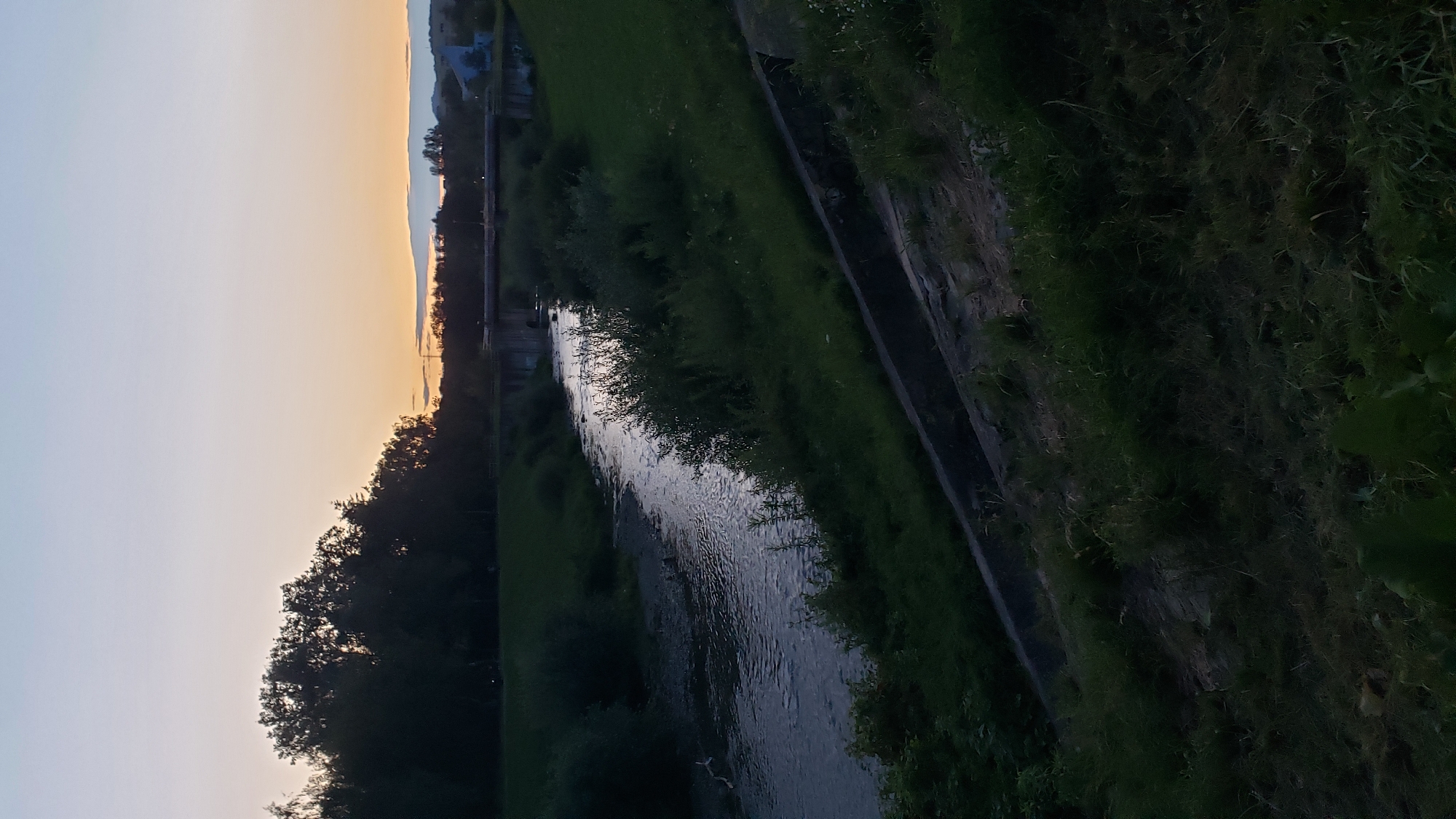 The image depicts a serene river flowing through a park-like setting at dusk. The river, with its gently rippling surface reflecting the fading light, meanders through a landscape characterized by lush green grass and dense trees lining its banks. In the distance, a bridge spans the river, hinting at the presence of infrastructure within the natural environment. The sky above is a gradient of soft blues and oranges, typical of a sunset, casting a tranquil ambiance over the scene. The overall impression is one of peaceful coexistence between nature and human elements, a common sight in urban green spaces. This scene is consistent with areas found along the Torysa river in Prešov, Slovakia, particularly within its recreational zones that feature walking paths, bridges, and tree-lined riverbanks, such as those found in the vicinity of the Sídlisko II or Sídlisko III neighborhoods.