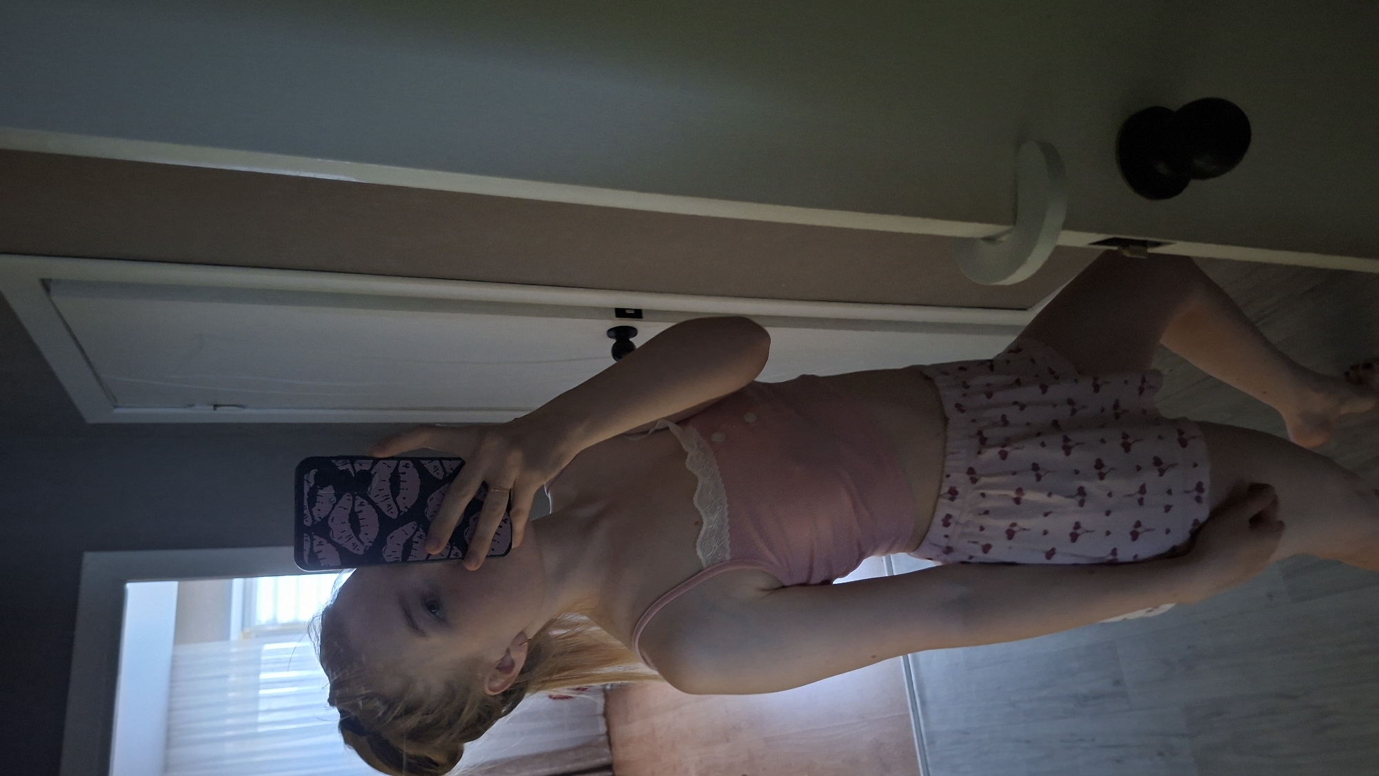 A young woman with blonde hair pulled into a bun is taking a selfie in a mirror. She is wearing a pink tank top with lace trim and pink shorts with a heart pattern. The background shows a room with a door and curtains. The image does not provide any information about the city of Chaikovsky, Russia.