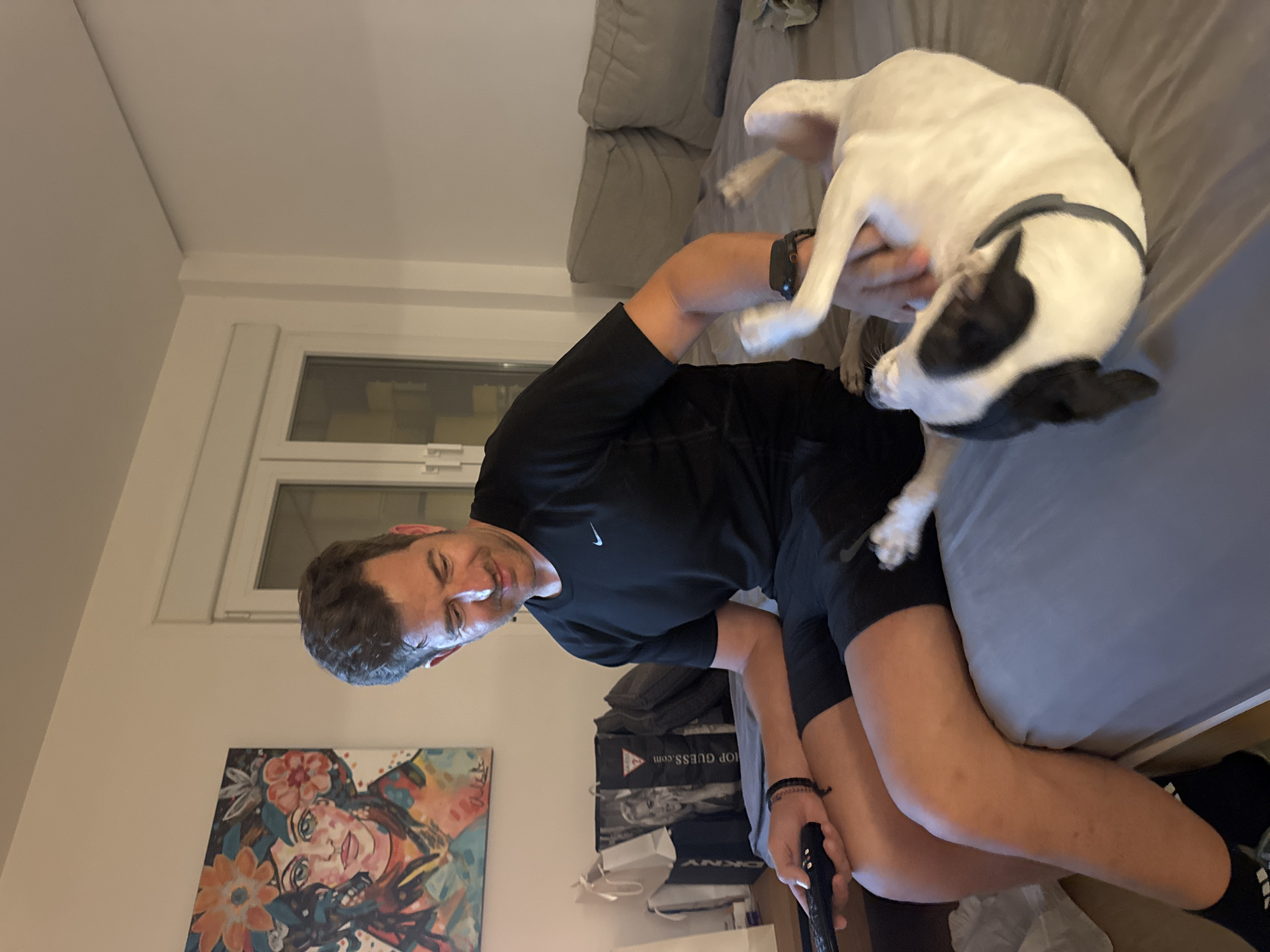 A man with short brown hair and a slight smile is sitting on a bed, holding a black remote control in his left hand. He is wearing a black Nike t-shirt and black shorts. His right hand is petting a white dog with black markings that is lying on its back on the bed, facing away from the camera. The dog appears to be enjoying the attention. The bed is made with grey linens and has several grey pillows at the head. Behind the man, to the left, is a framed painting of a woman's face adorned with flowers. There are also several shopping bags to the left of the painting, including one with a picture of a woman and the text "GUESS.COM". To the right of the man, a beige sofa is visible. The room is brightly lit by overhead lighting, and a window with white shutters is visible on the far right. The overall impression is a casual, domestic setting. The location is not discernable.