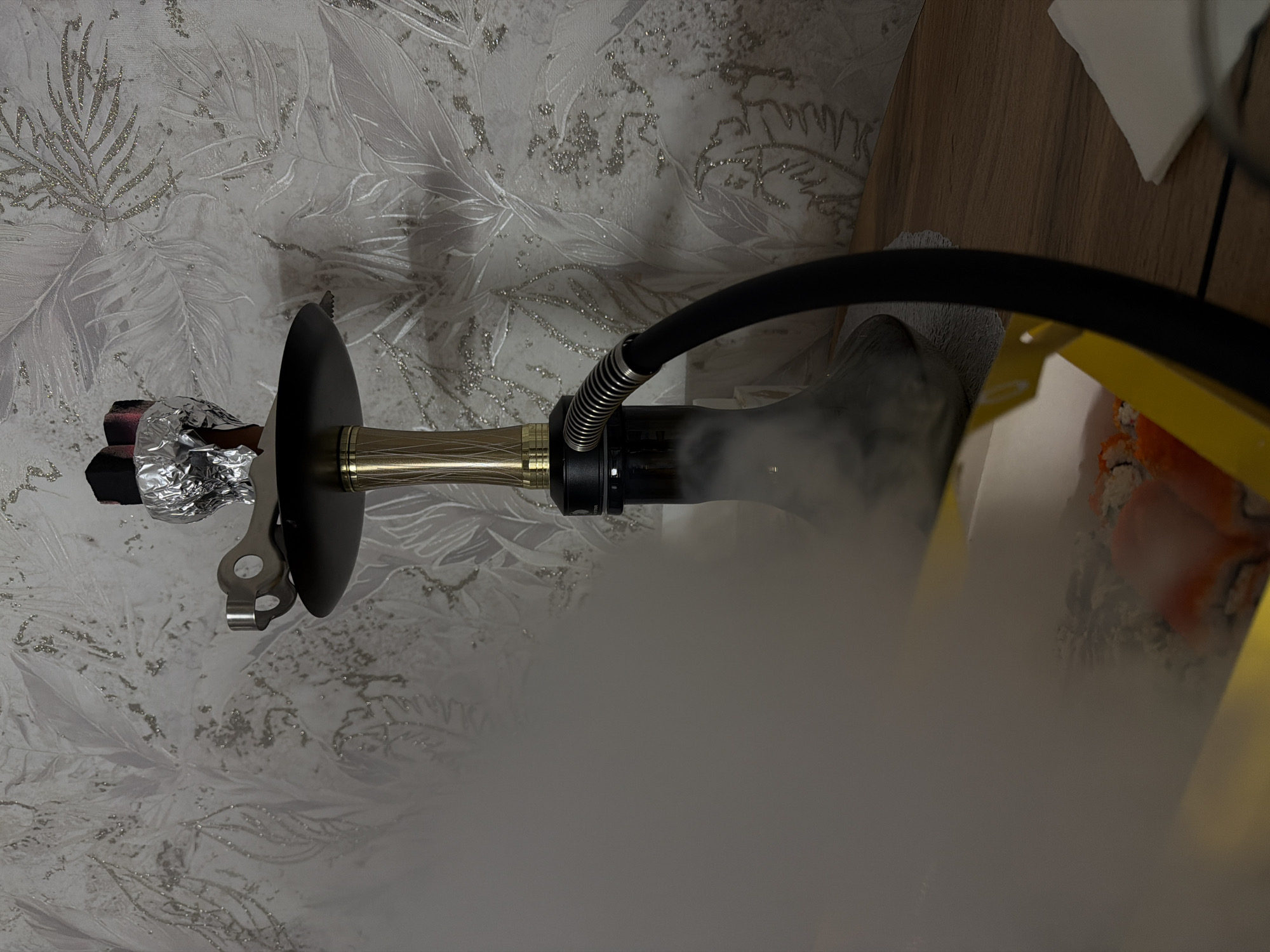 The image shows a hookah emitting smoke. The hookah consists of a foil-covered bowl, a black disc, a golden stem, a black hose with a coiled section, and a water-filled base. The background features a wall with a leaf-patterned wallpaper and a wooden surface with a white cloth and a yellow container holding sushi. There are no people visible. The image does not provide enough information to determine a specific location in Reshetnikovo, Russia.