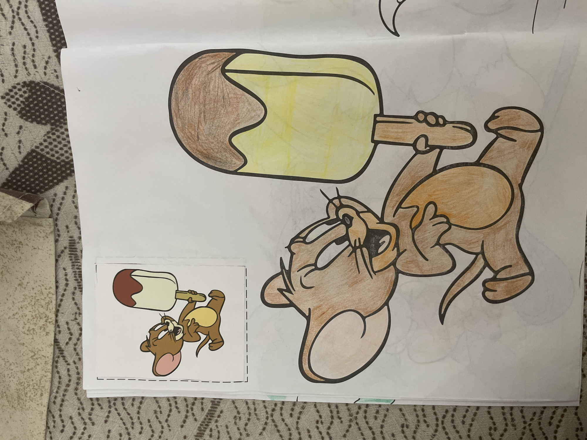 A partially colored drawing of Jerry from the animated series Tom and Jerry is featured on a white sheet of paper, placed on a patterned brown and beige textile surface, likely a rug or blanket. Jerry is depicted holding a large popsicle or ice cream bar, looking upwards with an open mouth, seemingly about to eat or enjoying the treat.
Jerry's body is colored in shades of brown, with a lighter brown or orange hue on his belly. His inner ears are pink, and his eyes are outlined in black. The popsicle stick is colored brown, while the ice cream portion is yellow with a brown, possibly chocolate, coating on one side.
In the bottom left corner, a smaller, printed and fully colored reference image of the same scene is visible. In this reference, Jerry is a more uniform brown with a light yellow belly and pink ears, and the popsicle is white with a darker reddish-brown coating.
The image shows an indoor setting, focusing on a creative activity, likely coloring. No text is visible in the frame.